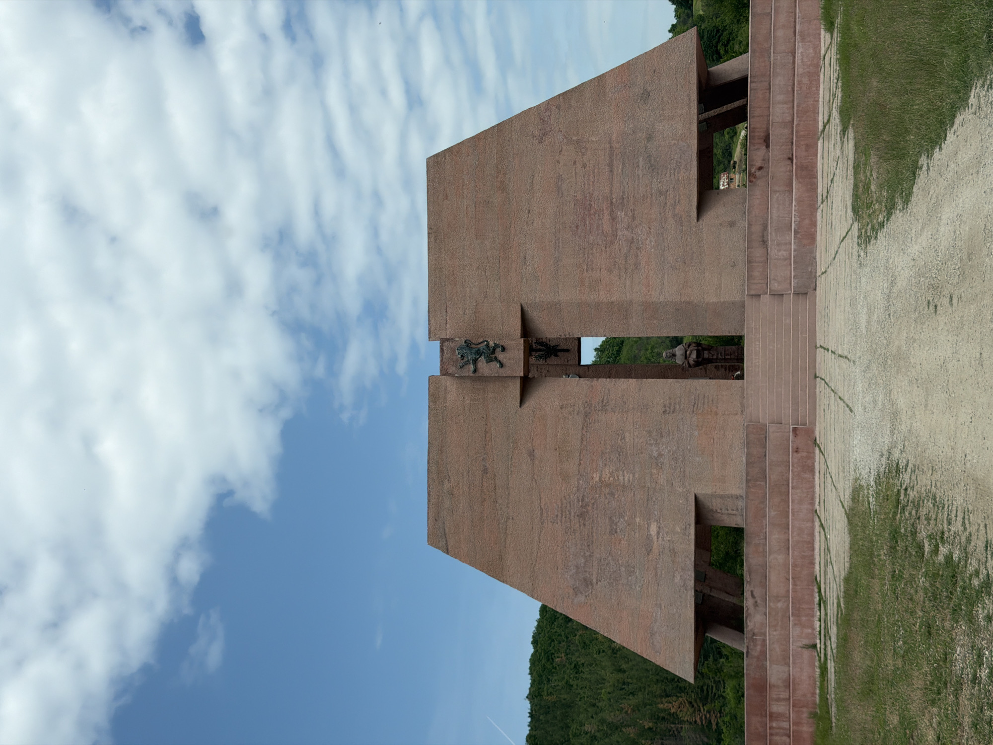 The image depicts the Monument "Chavdar Partisans" in Gurgulyat, Bulgaria. It is a large, reddish-brown concrete structure with several geometric forms. The main part of the monument features a prominent cross shape and a bas-relief depicting a lion, symbolizing strength and courage. The background includes a blue sky with scattered white clouds and green foliage from nearby trees. A pathway lined with grass leads to the monument. The structure has steps leading up to it. Through openings in the monument, we can glimpse more of the surrounding area. A statue of a person can be seen on the other side of the monument.
