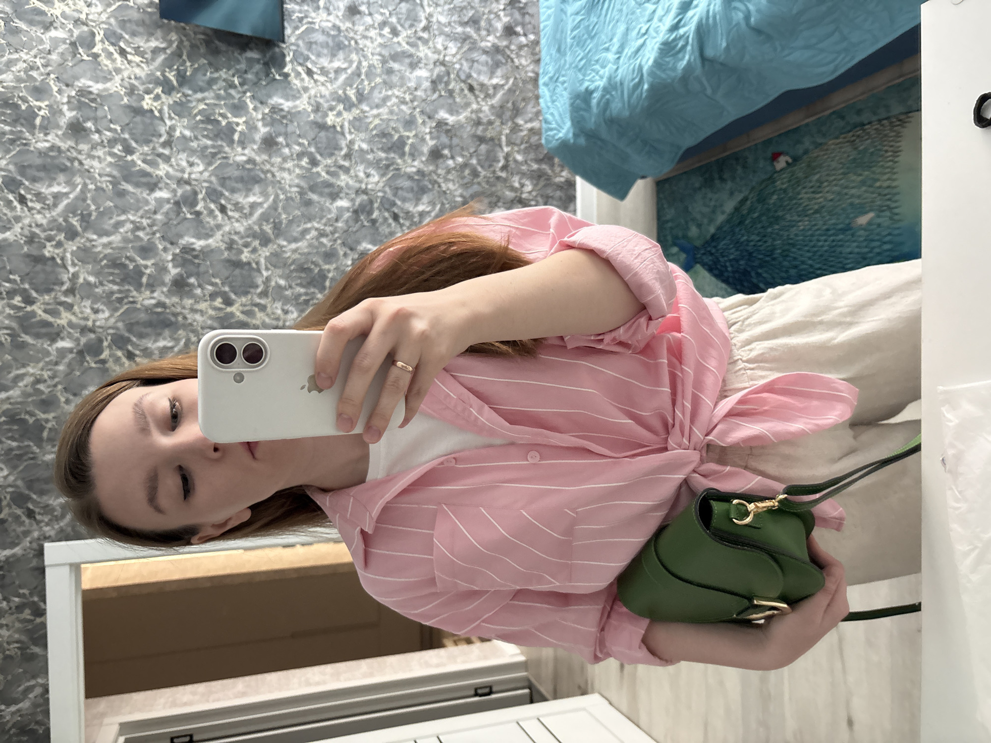 A young woman is taking a selfie in a bathroom mirror. She is wearing a white t-shirt, a pink and white striped button-down shirt tied at the waist, and light-colored pants. She is holding a white smartphone in her left hand. In her right hand, she holds a small green purse. The bathroom has a grey and white marble-like wall covering, a white cabinet, and a white door. A bed with blue bedding is visible in the background. The image was not taken in Izhevsk, Russia.