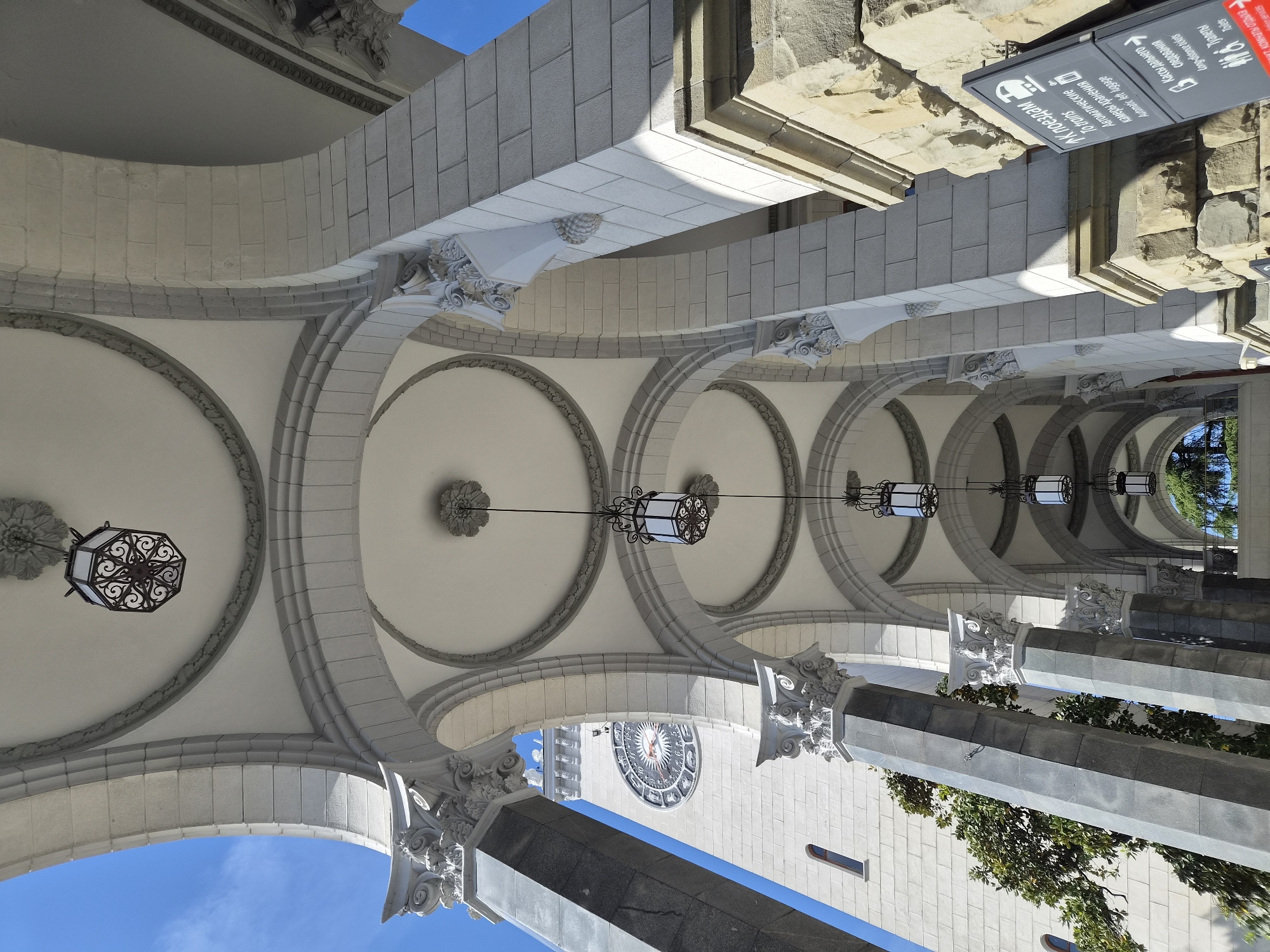 This image shows a view of an architectural structure in Sochi, Russia. The viewer is looking up at a series of repeating archways, each adorned with ornate decorative moldings and stone details. Suspended from the center of each archway is a pendant light fixture with a geometric design. The structure's design is a blend of classical and baroque elements. Between the archways, one can see sections of a building façade, including a clock tower with a large clock face. The sky is visible through openings in the structure. A sign post is visible that has some text and graphics in both English and Russian.