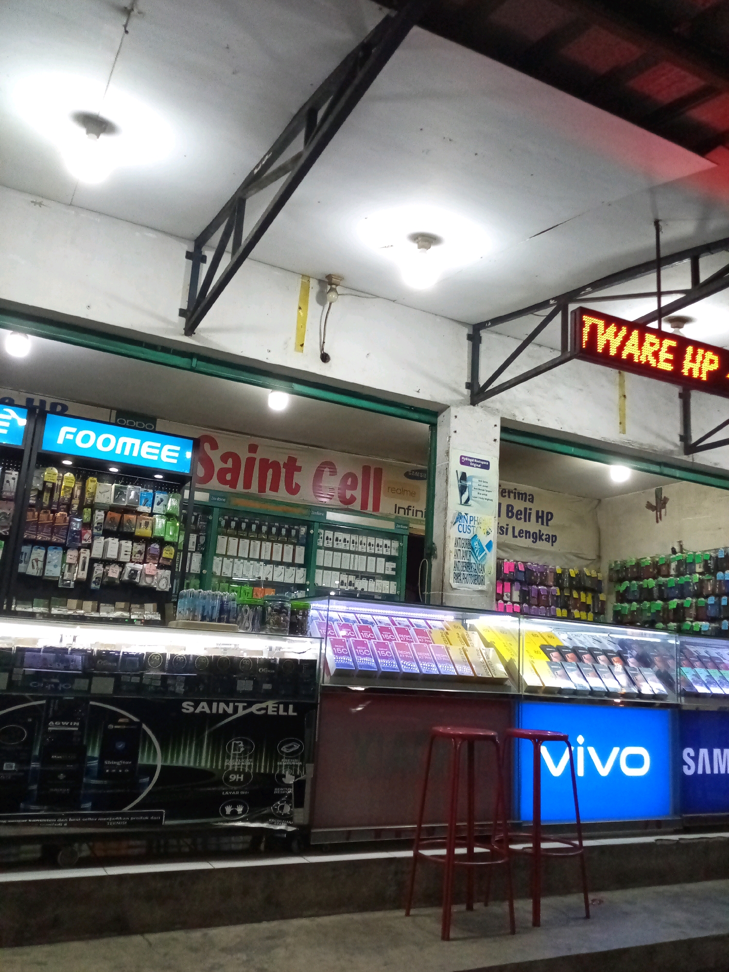A night time outdoor shot of a cell phone store named "Saint Cell" in Bekasi, Indonesia. The store is illuminated by bright lights, with a prominent "FOOMEE" sign and a red LED sign displaying "TWARE HP". Shelves are stocked with various phone accessories and brands like OPPO, Samsung, and Vivo. Two red bar stools are placed in front of the store. The overall scene depicts a typical retail environment for electronics in Indonesia.