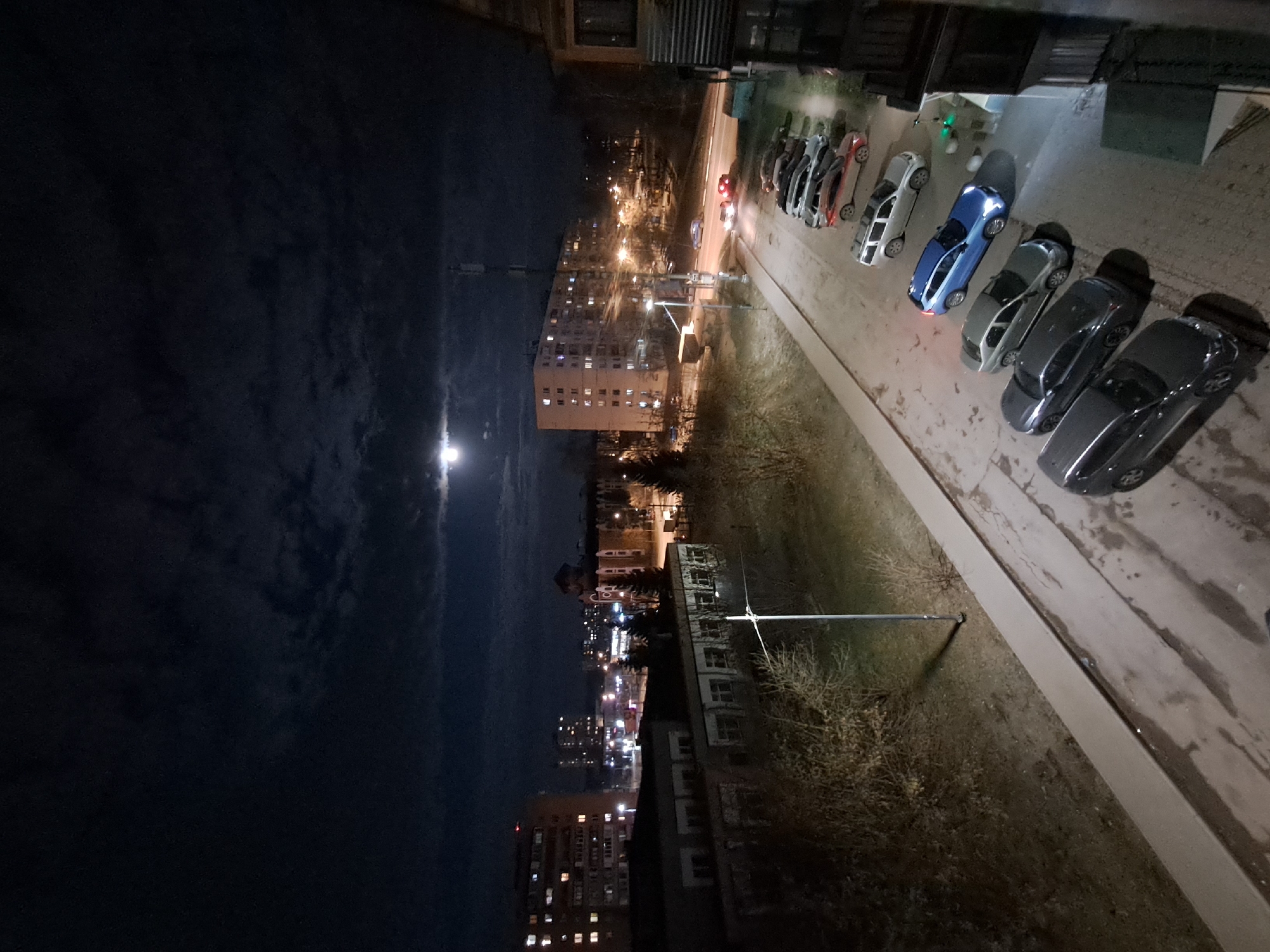 The image shows a nighttime view from a high vantage point, likely a balcony or window, in Elektrostal, Russia. The scene is dominated by a dark, cloudy sky with a visible moon. Below, the city is illuminated with streetlights and lights from buildings. A tall apartment building is prominent in the mid-ground, its windows glowing with light. A row of parked cars is visible along the right edge of the frame. The environment includes trees and other vegetation bordering the parking area. The scene lacks any people.