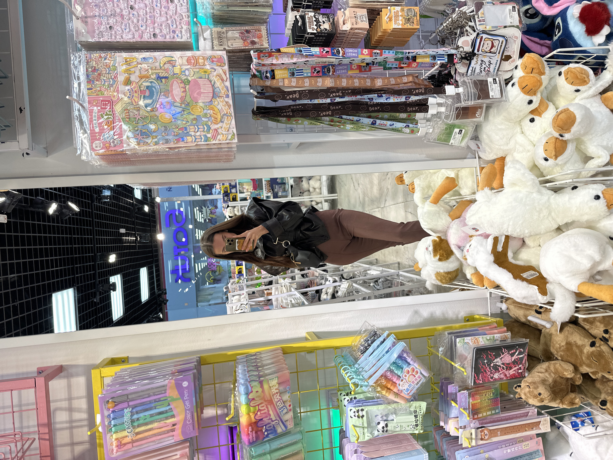 The image shows a reflection of a woman taking a picture with her phone in a store. The store appears to sell stationery, plush toys, and other novelty items. The reflection shows a grid-like ceiling with lights and a sign in Russian, indicating a location in Russia. Based on the sign, the store is located in Yekaterinburg. The woman is wearing a black jacket and brown pants. The shelves are stocked with items such as sticker sheets, lanyards, pens, plush ducks and teddy bears, and compass sets.