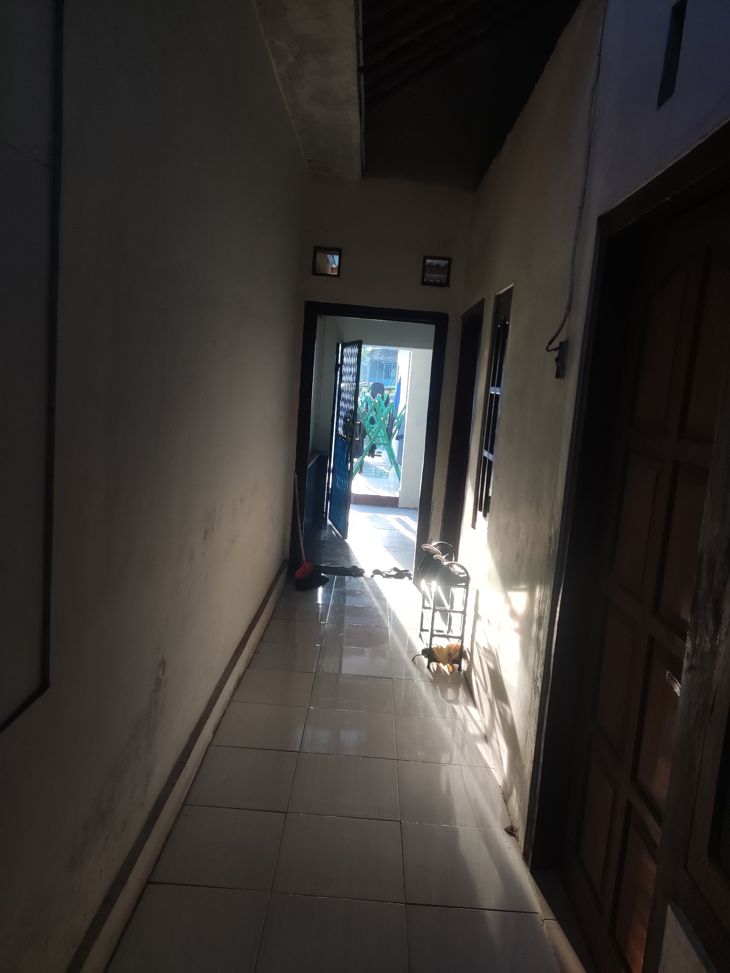 The image depicts a sunlit hallway in a residential building in Trihanggo, Indonesia. The hallway is long and narrow, with tiled flooring and cream-colored walls. Sunlight streams in from an open doorway at the far end, casting strong shadows across the floor and walls.
To the left, a plain wall extends down the length of the hallway. On the right side, there is a dark wooden door, partially visible and closed. Further down the hallway, there is another doorway with an open door, revealing a glimpse of an outdoor area with green barriers and sunlight. A broom with an orange handle leans against the wall near this doorway.
In the middle of the hallway, near the open doorway, a metal rack with several items is placed. These items appear to be light-colored, possibly bags or shoes, and are illuminated by the sunlight. The ceiling is partially visible at the top of the image, with exposed beams and a sloped roof. The overall impression is of a simple, functional hallway, possibly in a home or a modest commercial building, bathed in the strong light of the sun.
