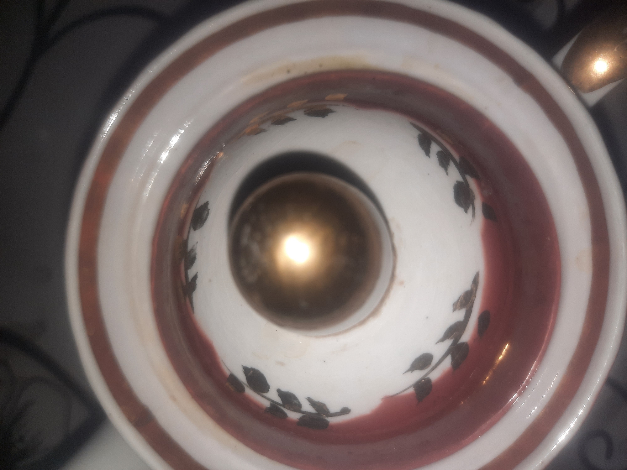 The image shows the top view of a ceramic object, possibly a teapot lid or a dish, with a central golden knob. The object has white and reddish-brown stripes running concentrically. There are also black decorative floral motifs on the white surface. The lighting suggests an indoor environment, but no specific location in Shklow, Belarus can be determined from the image alone.
