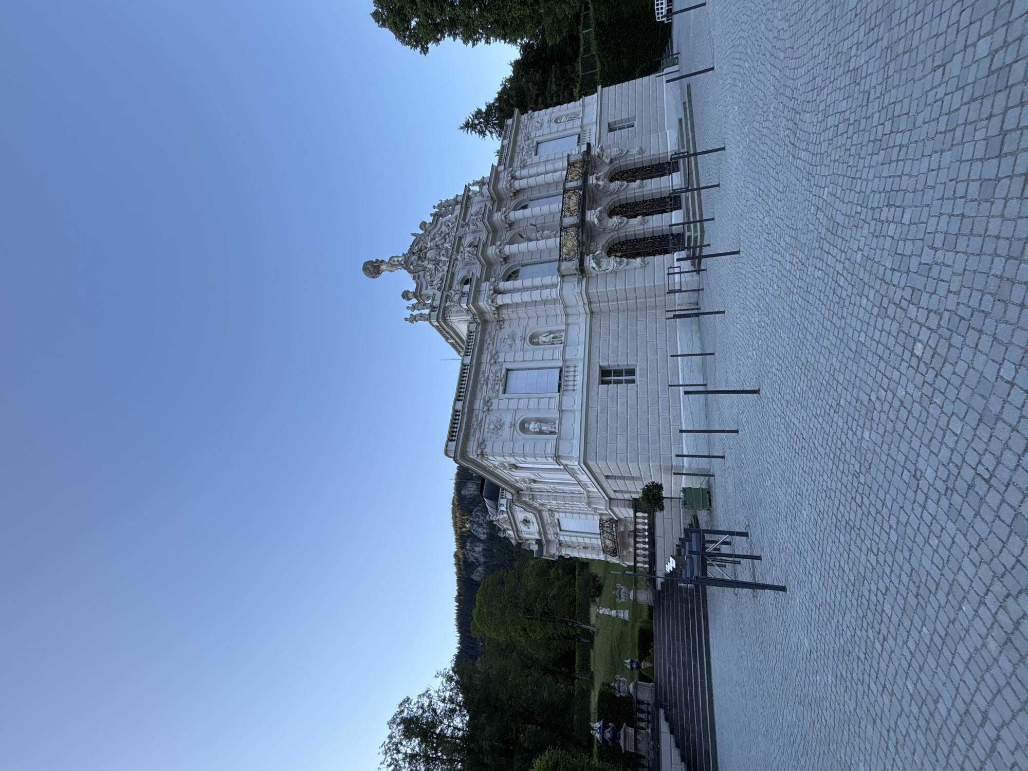 A bright, clear day in Ettal, Germany, shows Linderhof Palace, a royal villa with ornate white stucco and grey stone construction. The palace features multiple balconies, columns, and intricate statues adorning its facade. In front of the palace is a large, cobblestone plaza with several low, black metal stanchions. To the left of the palace, a grand stone staircase leads up to a manicured lawn dotted with more statues and urns. Beyond the lawn, lush green trees cover a steep hillside that forms the backdrop. The sky above is a clear, pale blue, with no visible clouds. The sun casts soft shadows, indicating it is likely late afternoon or early morning.