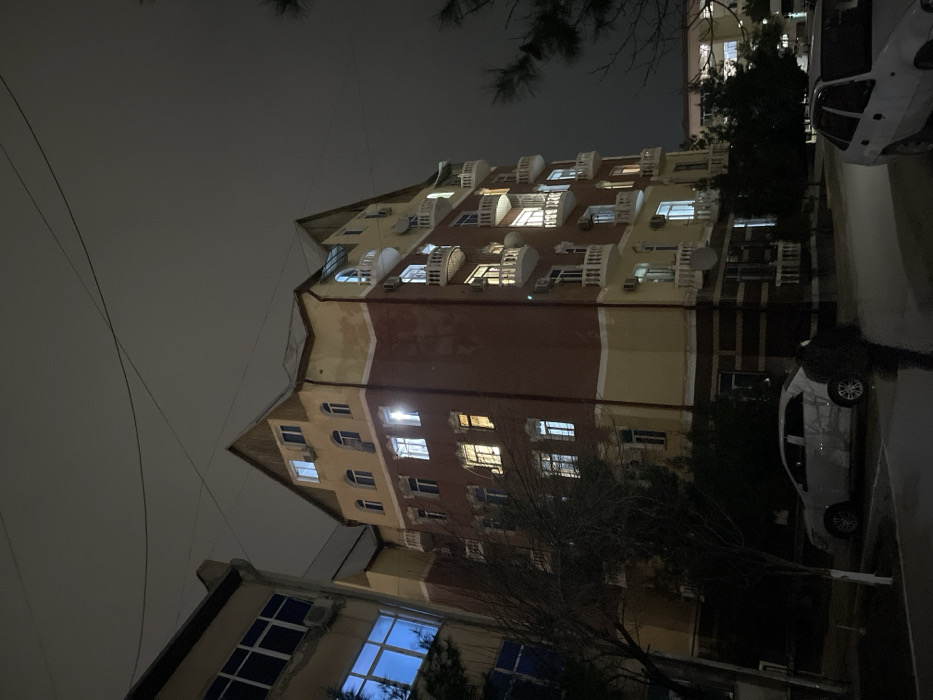 This nighttime photograph captures a street scene in Navoiy City, Uzbekistan. The central focus is a multi-story residential building, approximately six floors in height, featuring a gabled roof section and a facade segmented by color: a reddish-brown middle section flanked by lighter yellow or beige upper and lower portions, accented with white trim. Numerous windows are visible, many illuminated with either warm yellow or cool white light. The building displays multiple white, curved balconies and external air conditioning units, along with at least two white satellite dishes mounted on its exterior.
To the left of the central building, a section of an adjacent multi-story structure is visible, characterized by large, dark-paned windows, some reflecting a distinct blue light, and an external air conditioning unit. The sky above is dark and overcast, with several power lines traversing the upper frame.
At ground level, a paved area or sidewalk extends across the foreground. A lone individual, dressed in dark clothing, is observed walking away from the camera in the lower center-right of the frame. Two sedans are parked: a light-colored vehicle is positioned on the left, facing right, and a white sedan is partially visible on the far right, facing left. Sparse, bare-branched trees and some coniferous trees line the street and are situated near the buildings.