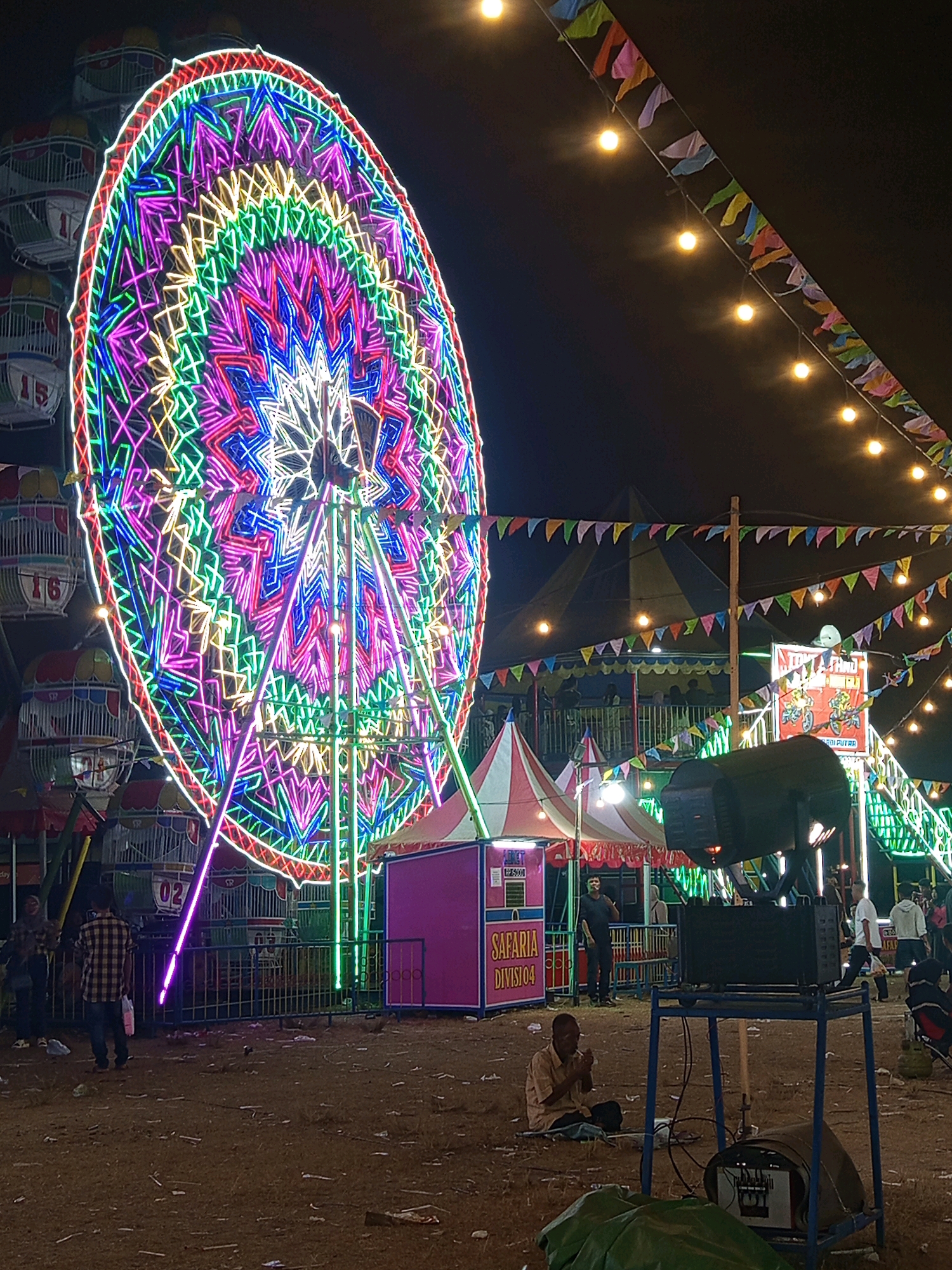 This image depicts an outdoor nighttime scene at a carnival or fairground in Pulung Kencana, Indonesia. The dominant feature is a brightly illuminated Ferris wheel adorned with a complex pattern of colorful LED lights in shades of pink, purple, blue, green, and yellow. Several gondolas on the Ferris wheel are visible, with numbers like "15" and "16" on some of them. In the foreground, a man is seated on the ground, appearing to be praying or meditating. Adjacent to him is a blue metal stand holding audio equipment, including a large black speaker and a mixer. The ground is covered with dirt and scattered debris. To the right of the Ferris wheel, there is a smaller tent with "SAFARIA DIVISI 04" written on it. Further in the background, other carnival attractions are visible, including what appears to be a smaller ride or game stall with a tent-like structure. String lights with small colorful flags are draped across the scene, adding to the festive atmosphere. Several people are present in the background, some standing near the rides and others walking. The overall impression is a lively, illuminated carnival at night.