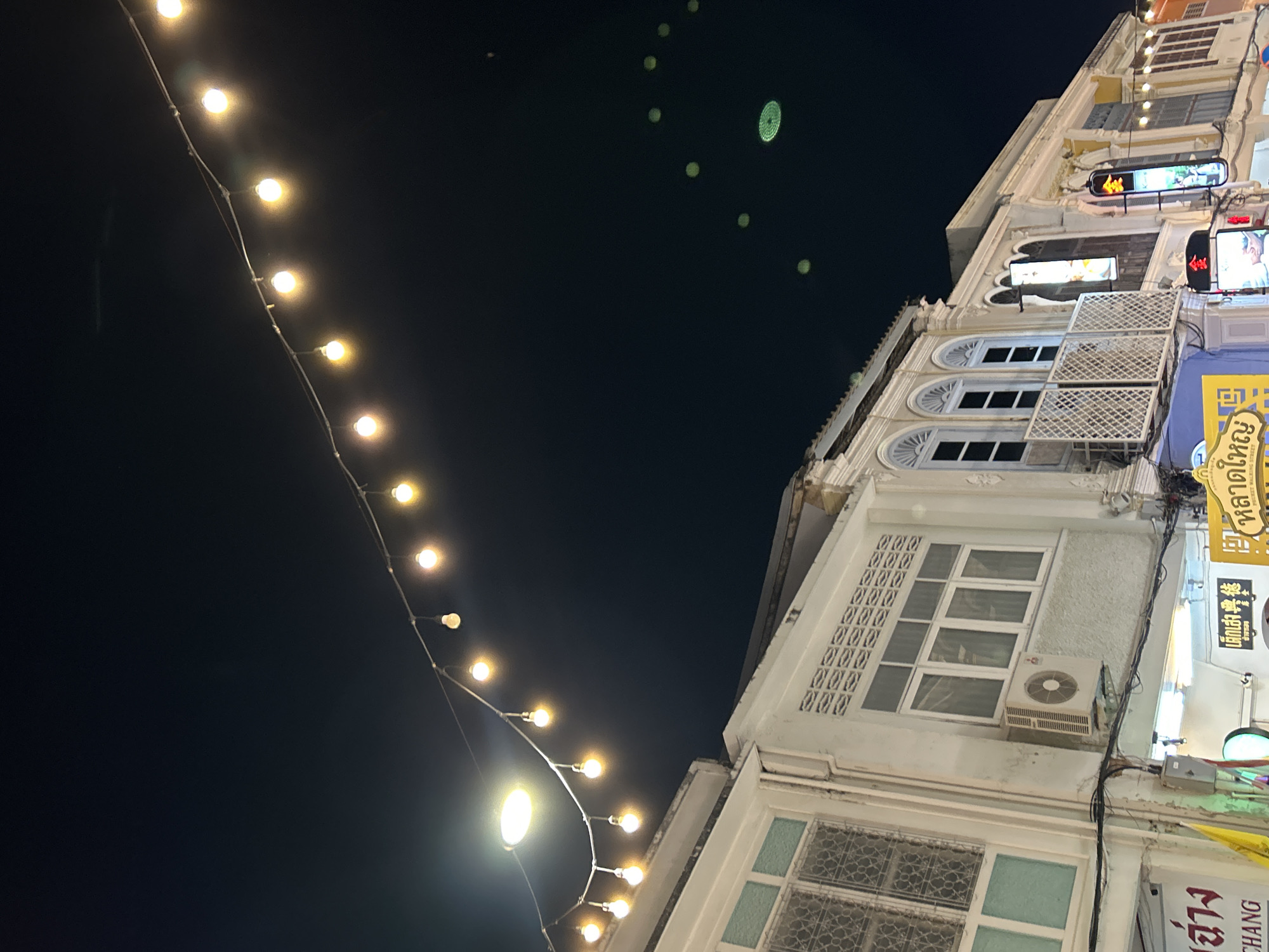 An outdoor, eye-level, medium shot captures the facade of several historical Sino-Portuguese buildings at night. The buildings are illuminated by a string of warm, glowing lightbulbs suspended across the dark sky. The architecture features white and cream-colored walls with decorative window frames, balconies with intricate metalwork, and louvered vents. An air conditioning unit is mounted on one of the walls. Below the buildings, there are visible signs with Thai script. In the upper right quadrant of the image, there are several small, round, green lights visible against the dark sky. The image appears to be taken on Soi Romanee, a famous street in Phuket City Municipality, Thailand, known for its well-preserved architecture and vibrant atmosphere, particularly at night. No people are visible in the scene.