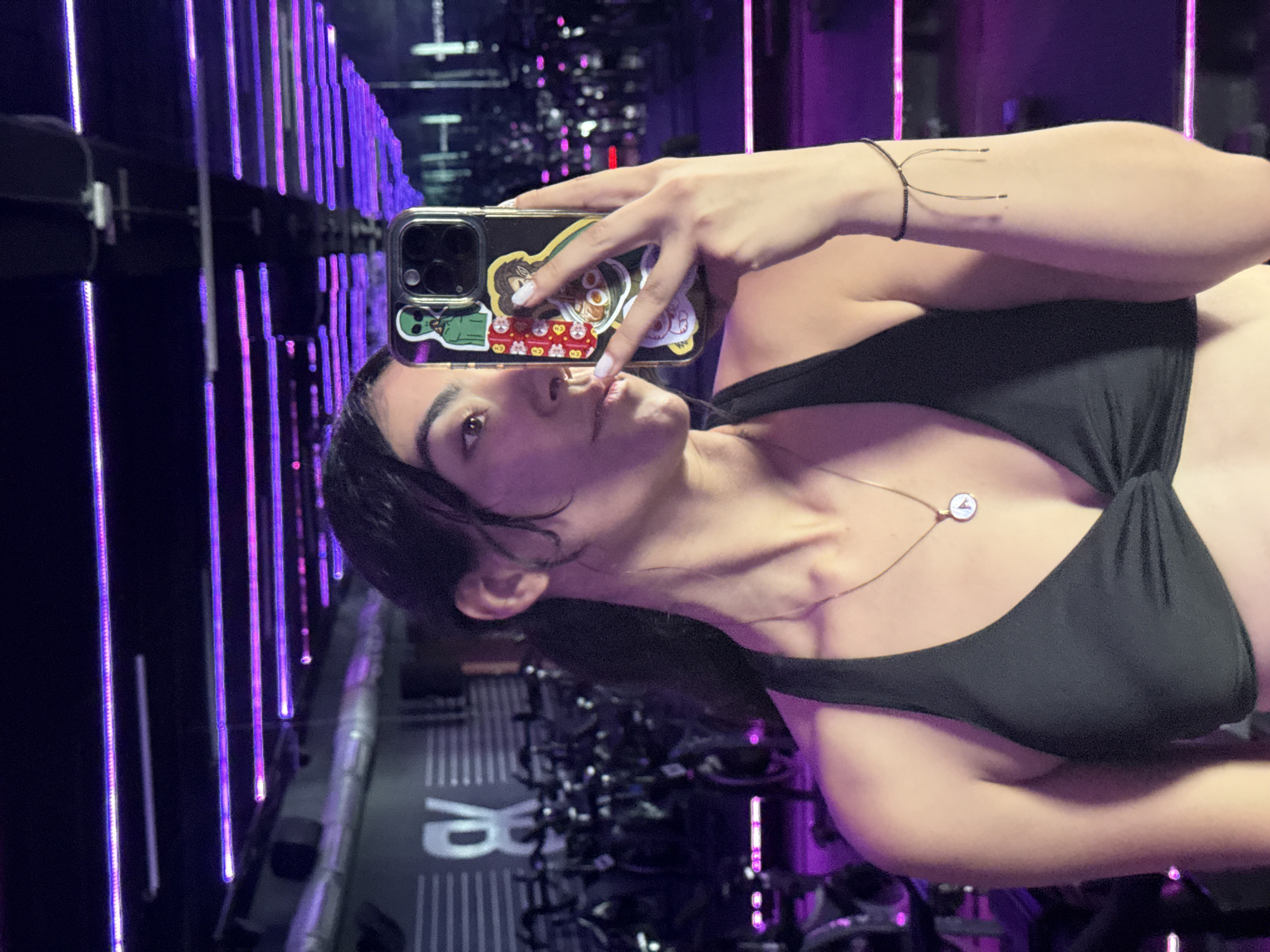 The image shows a person taking a selfie in what appears to be a gym setting. The individual has dark hair and is wearing a black top with a gold necklace. They are holding a smartphone with stickers on its case. Behind the person, there are rows of stationary bikes and vertical purple light fixtures. The gym name 'RB' is visible in large letters on the floor. There is no evidence to confirm the location in the city of Puebla, Mexico.
