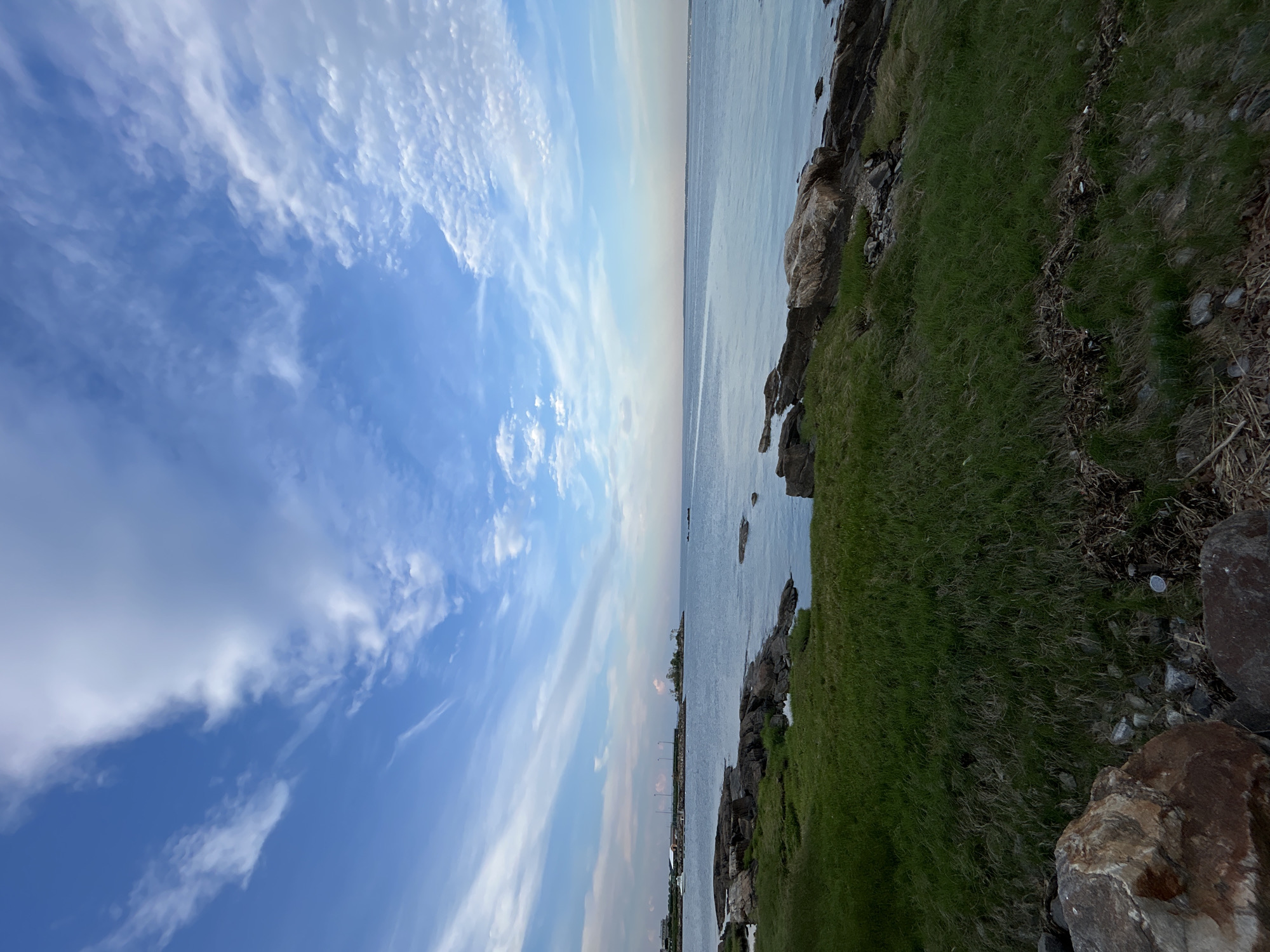 The image shows a landscape in New Rochelle, United States of America. On the right, there is a grassy area with rocks. The horizon line separates the grass from the ocean, which extends towards the center of the image. Above the ocean is the sky, which features a combination of blue patches and streaks of white clouds. The image appears to be taken at either sunrise or sunset, as the light seems soft and diffused. The composition is vertically oriented.