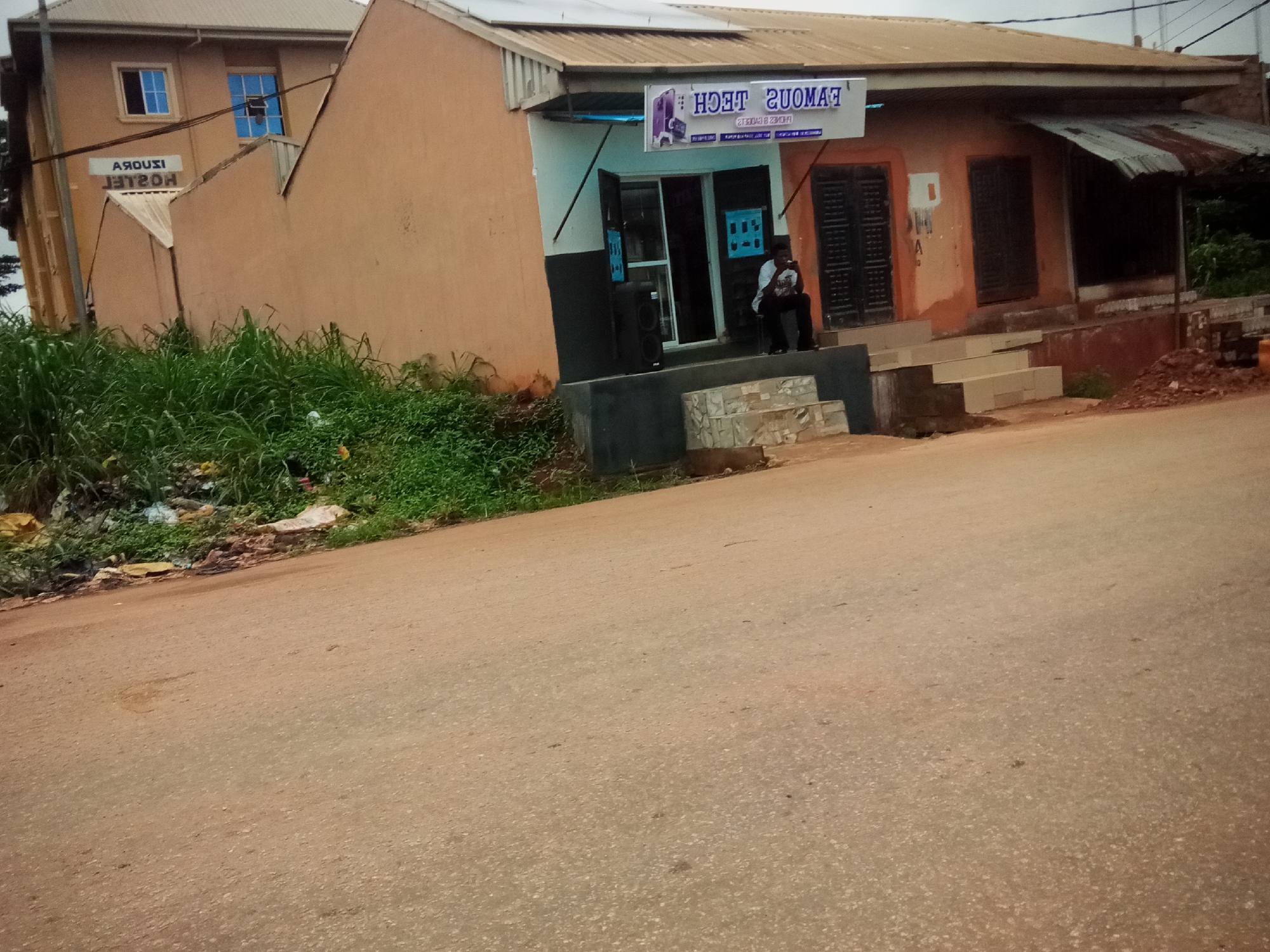 The image depicts a scene in front of a small commercial establishment and a hostel in Oko Town, Nigeria. The commercial building, identified as "FAMOUS TECH," features a sign with purple lettering and an image of a smartphone. The storefront is open, and a person is seated on the steps outside. Next to the storefront, a large black speaker is visible. The hostel, labeled "IROUZI HOSTEL" in white lettering on a dark background, is a multi-story building with tan walls and blue-framed windows. Adjacent to the buildings, there is overgrown green grass and accumulated debris, including plastic bags. The foreground shows a paved road, likely a street, with some discoloration and minor debris. The environment appears to be an urban or semi-urban setting.