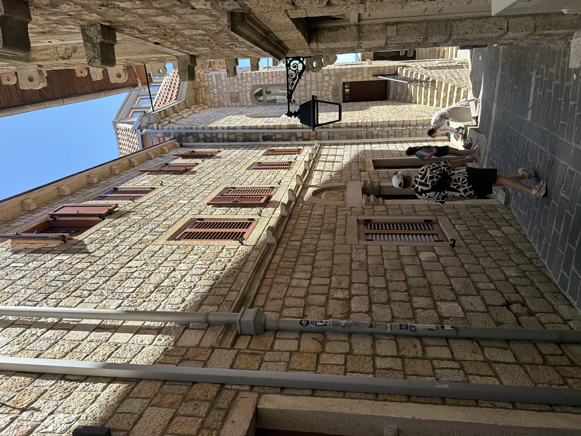 The image depicts a narrow, cobblestone street in the old town of Ulcinj, Montenegro. The street is lined with traditional stone buildings, characterized by their rough-hewn blocks and dark wooden shutters. A tall, grey downspout runs vertically along the left-hand building. In the mid-ground, a black wrought-iron lantern hangs from the side of a building. A set of stone stairs with a metal railing leads upwards to a wooden door.
Several individuals are present in the scene. In the foreground, a person wearing a black and white patterned shirt, dark shorts, a white cap, and sneakers walks away from the camera. Further down the street, two young women stand near the stairs. One is wearing a blue top and a dark skirt, while the other is wearing a light-colored outfit and appears to be bending down to retrieve an item. The bright sunlight casts sharp shadows across the street and buildings, suggesting a clear, sunny day. The sky is a vibrant blue. The overall impression is of a well-preserved historic urban environment.