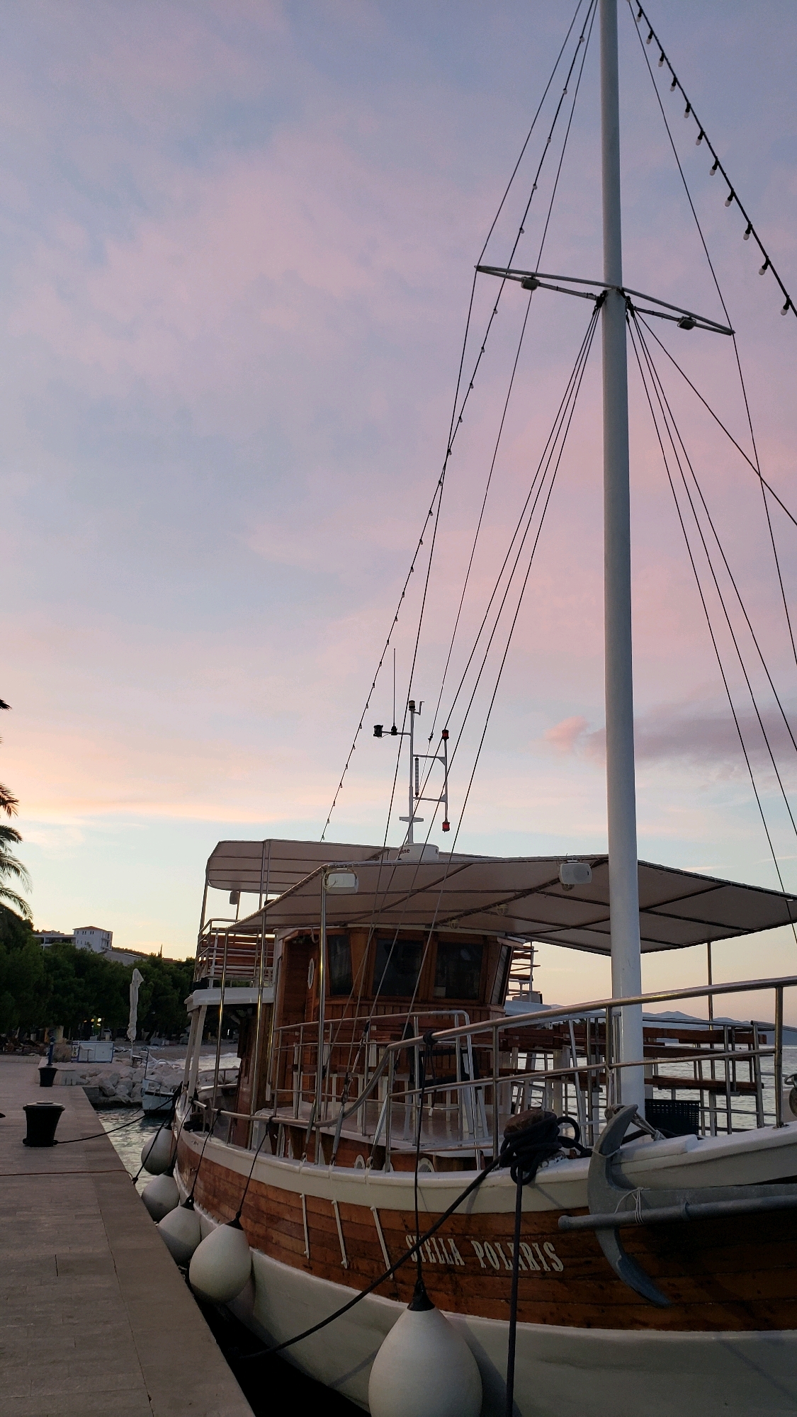 The image shows a wooden boat named "STELLA POLARIS" docked at a harbor in Općina Tučepi, Croatia, during a sunset. The sky displays soft pastel hues of pink and blue. The boat is prominently featured in the foreground, with its wooden hull and white trim visible. White fenders are attached along its side, hanging from black ropes. The boat has multiple decks with railings and covered seating areas. A tall mast with rigging and lights is also visible. In the background, there is a stone walkway, lush green trees, some buildings on a hill, and other boats in the water. The overall scene is serene and picturesque, capturing the tranquility of the coastal town at dusk.