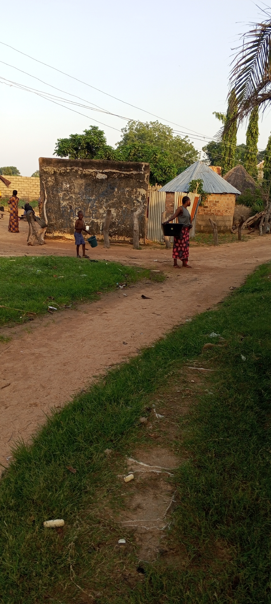 The image depicts a scene in Jalingo, Nigeria, characterized by a dirt road winding through a village. Several individuals are present, engaged in daily activities. A young boy stands near a textured wall, holding a blue bucket and what appears to be a cup or smaller container. To his right, a woman is seen walking with a large black basin on her shoulder. Further in the background, another woman in a patterned dress stands near a low wall. The environment features rudimentary structures, including a building with a corrugated metal roof and circular thatched huts, along with a large, weathered concrete structure. Vegetation, including grass and trees, is visible throughout the scene, interspersed with electrical wires overhead. The ground is a mix of dirt and patches of grass, with some scattered debris. The overall impression is one of a rural or semi-urban setting in Nigeria, capturing a moment of everyday life.