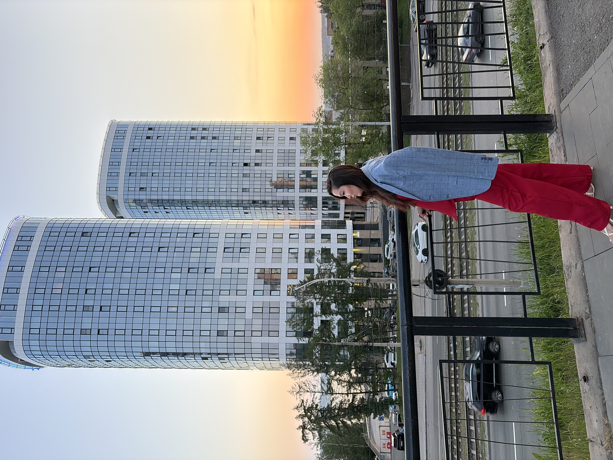 A full body shot shows a woman wearing a blue blazer and red pants. The woman is leaning over a black metal railing. Two modern curved high rise buildings are behind her. The buildings have many windows. A sunset is in the background. The scene is in Izhevsk, Russia. The photograph also shows a road behind the railing with multiple cars.