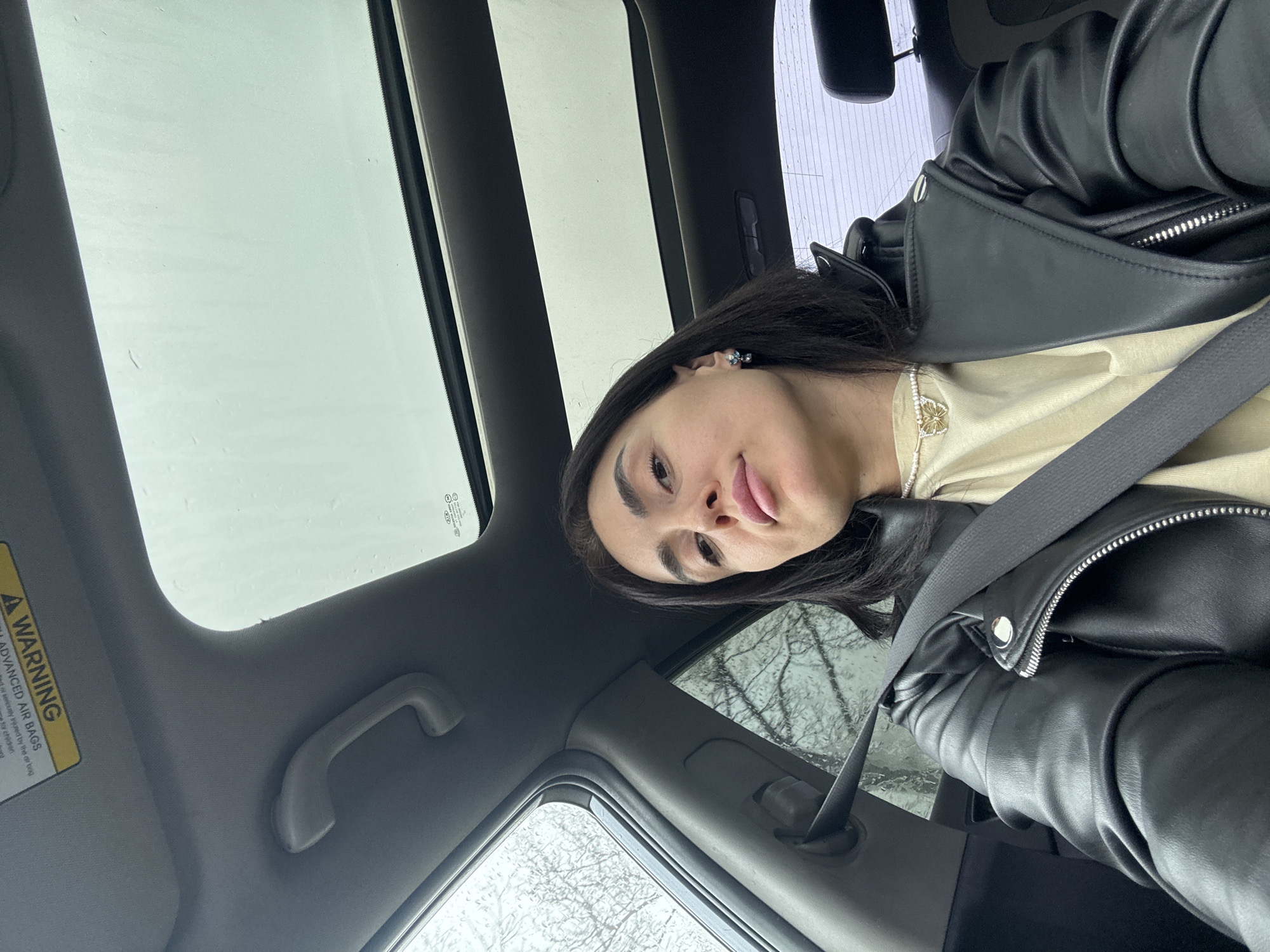 A female individual is depicted in a selfie taken from the front passenger seat of a Kia vehicle within the city of Tbilisi, Georgia. She has dark hair and is wearing a black leather jacket over a light tan t-shirt, secured by a dark seatbelt across her chest. A pearl necklace with a gold butterfly pendant is visible, along with a light-colored gemstone earring on her right ear. Her expression is neutral, directed towards the camera. The vehicle's interior features a gray headliner, a passenger grab handle, and a "WARNING ADVANCED AIR BAGS" label on the overhead visor. A sunroof shows an overcast sky with visible moisture. Through the side window on the subject's left, bare tree branches are observed against a gray sky, with water droplets on the glass, indicative of rain or high humidity. The background through the rear window is indistinct due to blur.