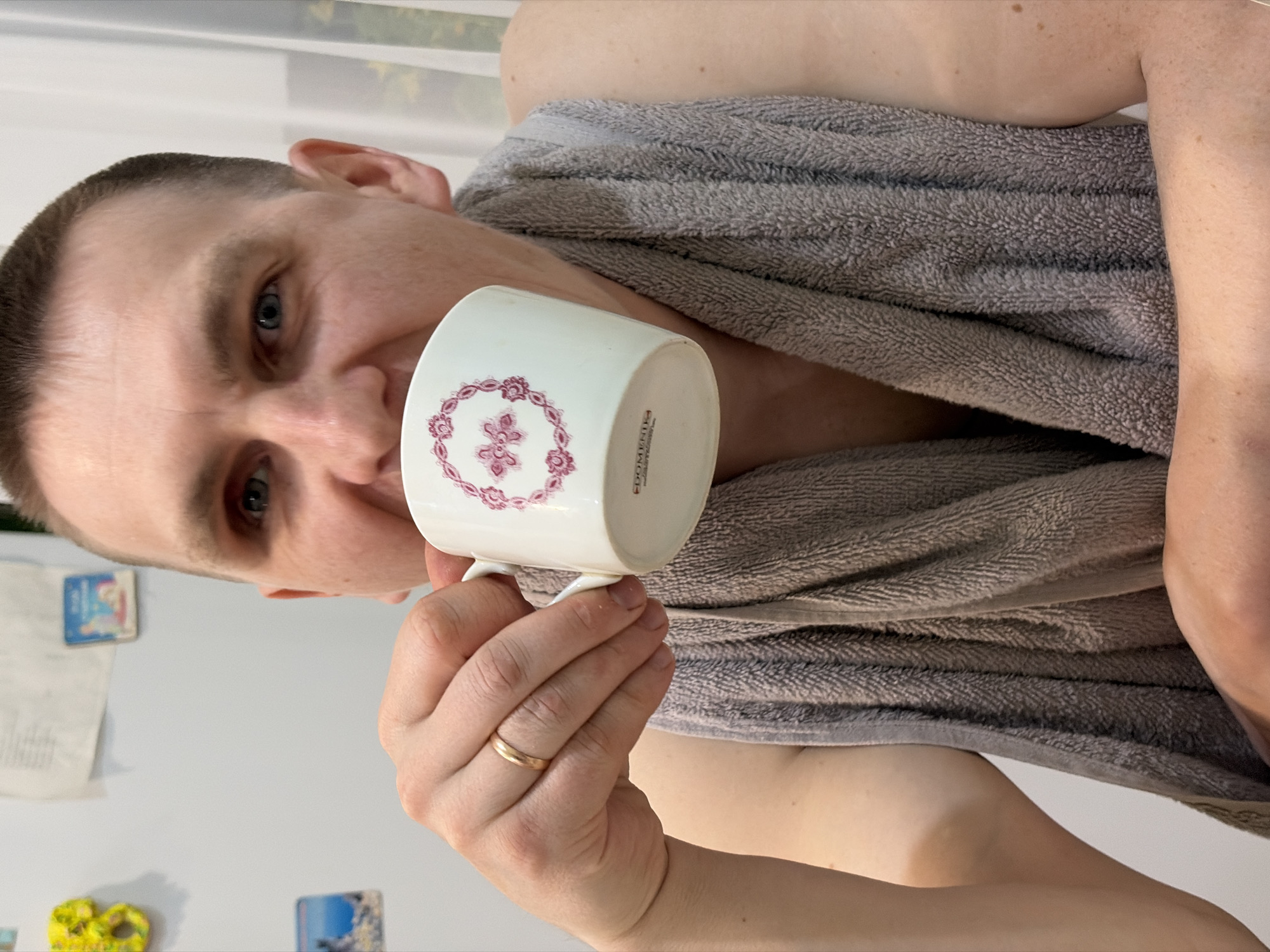 A man with a shaved head and blue eyes holds a white mug with a pink floral design. He wears a gray towel draped around his shoulders. A gold wedding band is visible on his left ring finger. In the background, a white refrigerator door is adorned with papers, a yellow pretzel magnet, and a blue magnet featuring a mountain landscape. The location of Noviy, Russia is not determinable from the image.