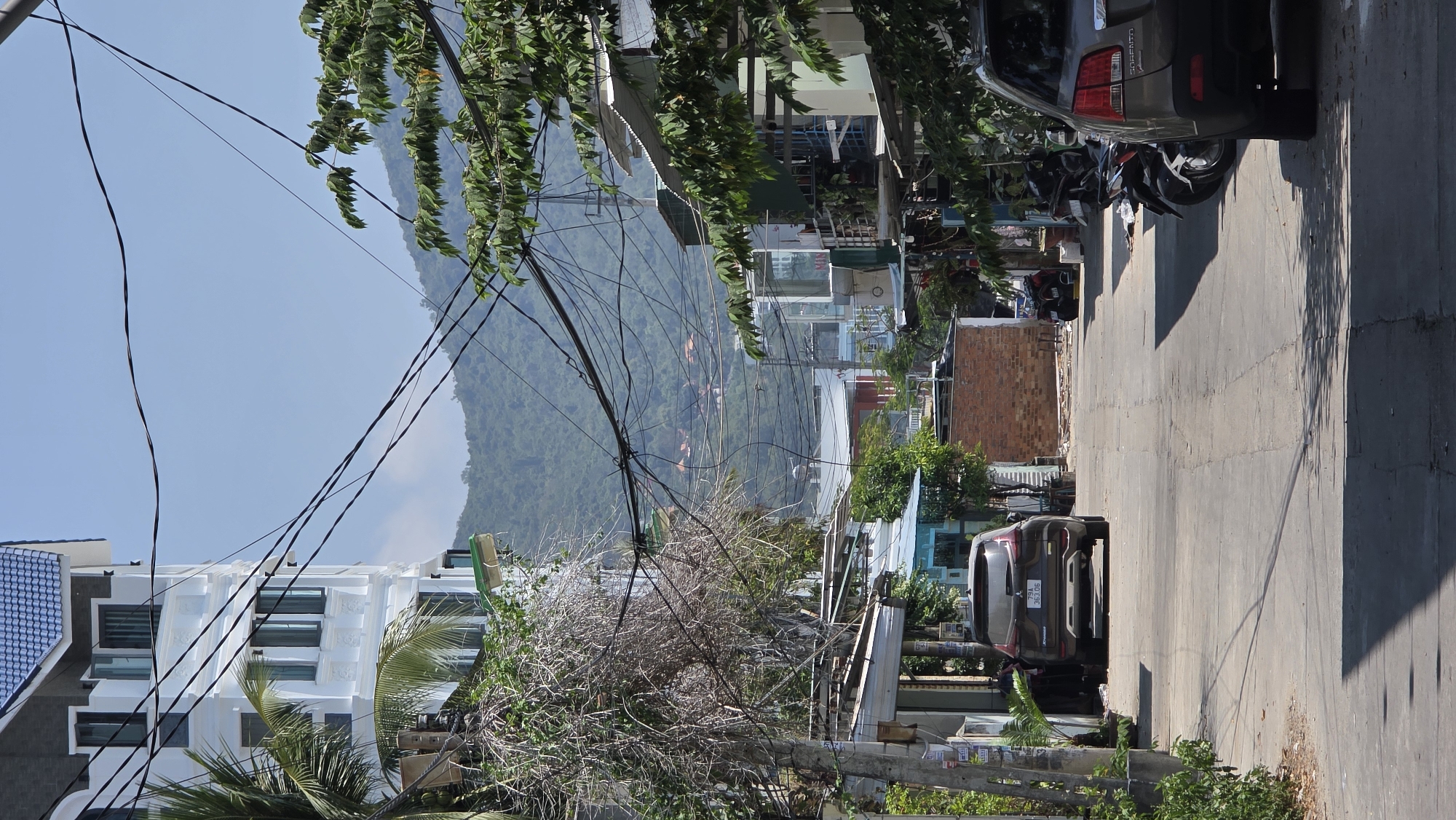 The image depicts a narrow street scene in the city of Bắc Nha Trang, Vietnam, seen from a slightly elevated perspective. The street runs vertically, with buildings lining both sides. The building on the left is white with a blue tiled roof. On the right is a mixture of commercial establishments with awnings. Utility poles are visible, strung with a multitude of overhead power lines that crisscross the sky. A dense, green mountain rises in the distance, partially obscured by the power lines and vegetation. There are parked cars and motorbikes along the edge of the road. The pavement is a light grey color and has visible cracks and imperfections. The lighting suggests a bright, sunny day.