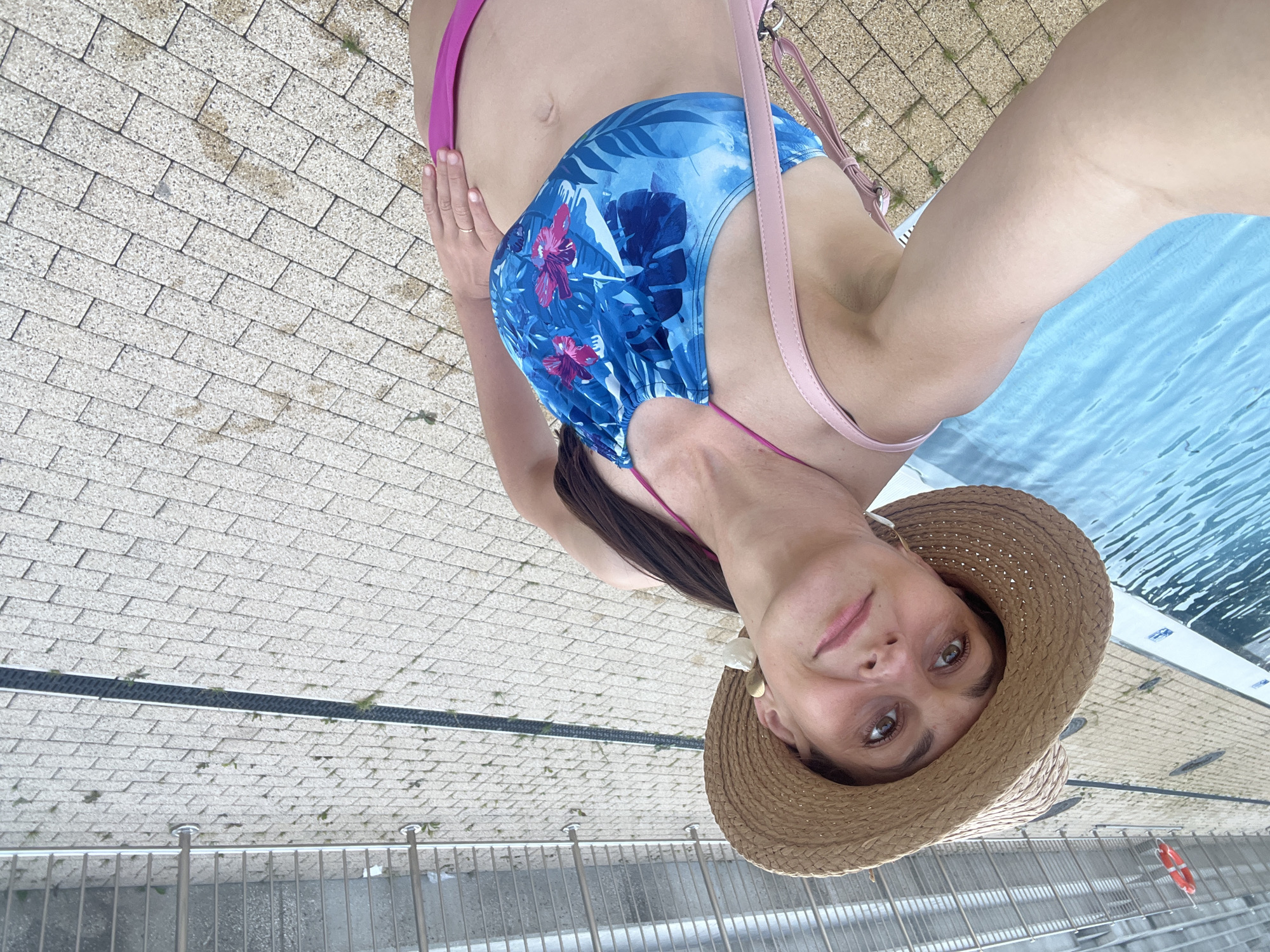 A woman is taking a selfie by a swimming pool. She is wearing a straw hat, a blue bikini top with a floral pattern, and pink bikini bottoms. A pink strap from a bag is visible over her shoulder. The woman is standing on a paved area next to the pool. In the background, there is a metal fence and a red life preserver. The water in the pool is blue.