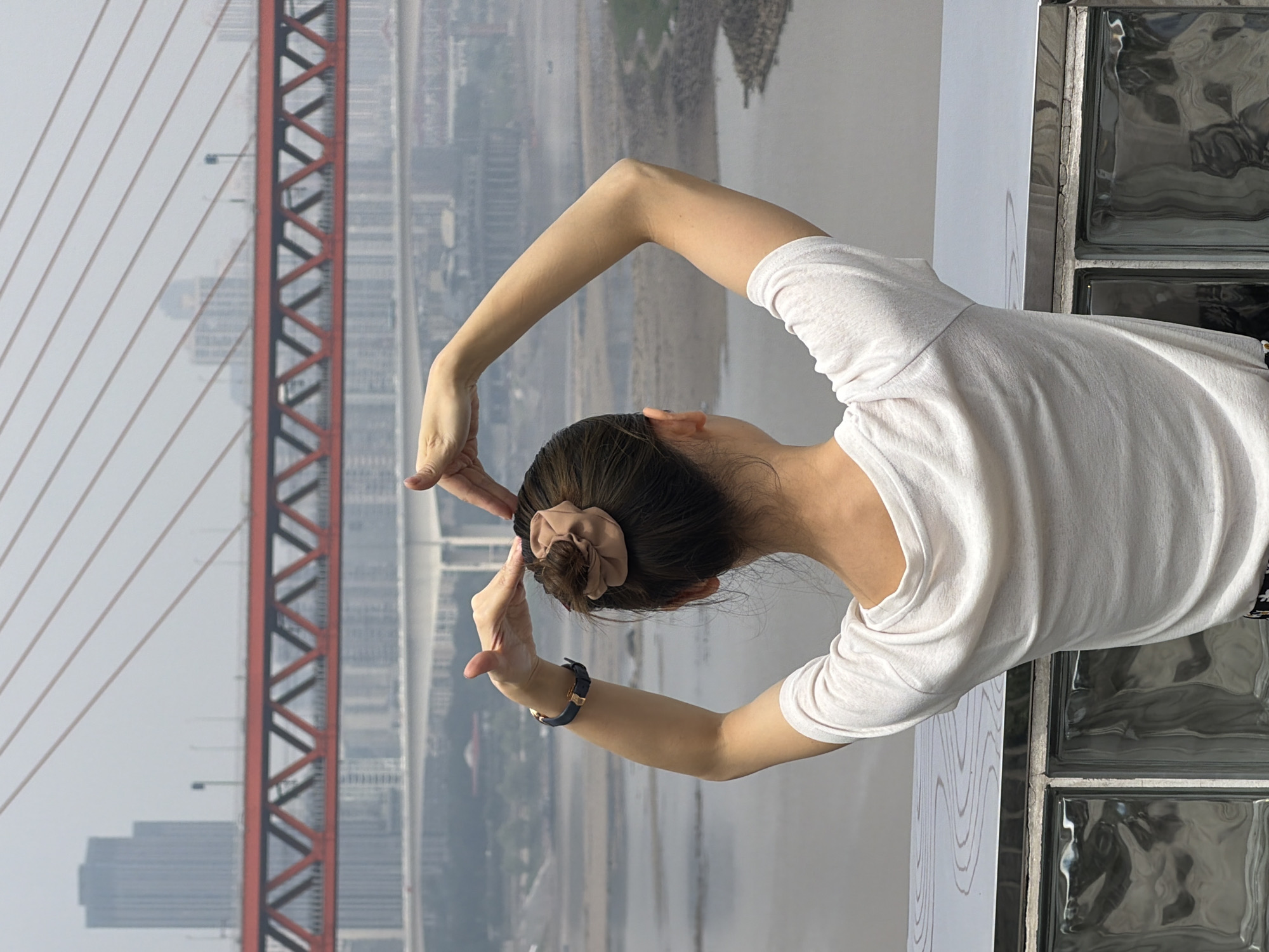 A person stands with their back to the camera, arms raised, forming a heart shape with their hands above their head. The person is wearing a white t-shirt and a scrunchie in their hair. They are positioned on a platform overlooking a body of water, with a red suspension bridge and city skyline in the background. This scene is located in Yuzhong District, China. The environment appears hazy and the day is overcast.