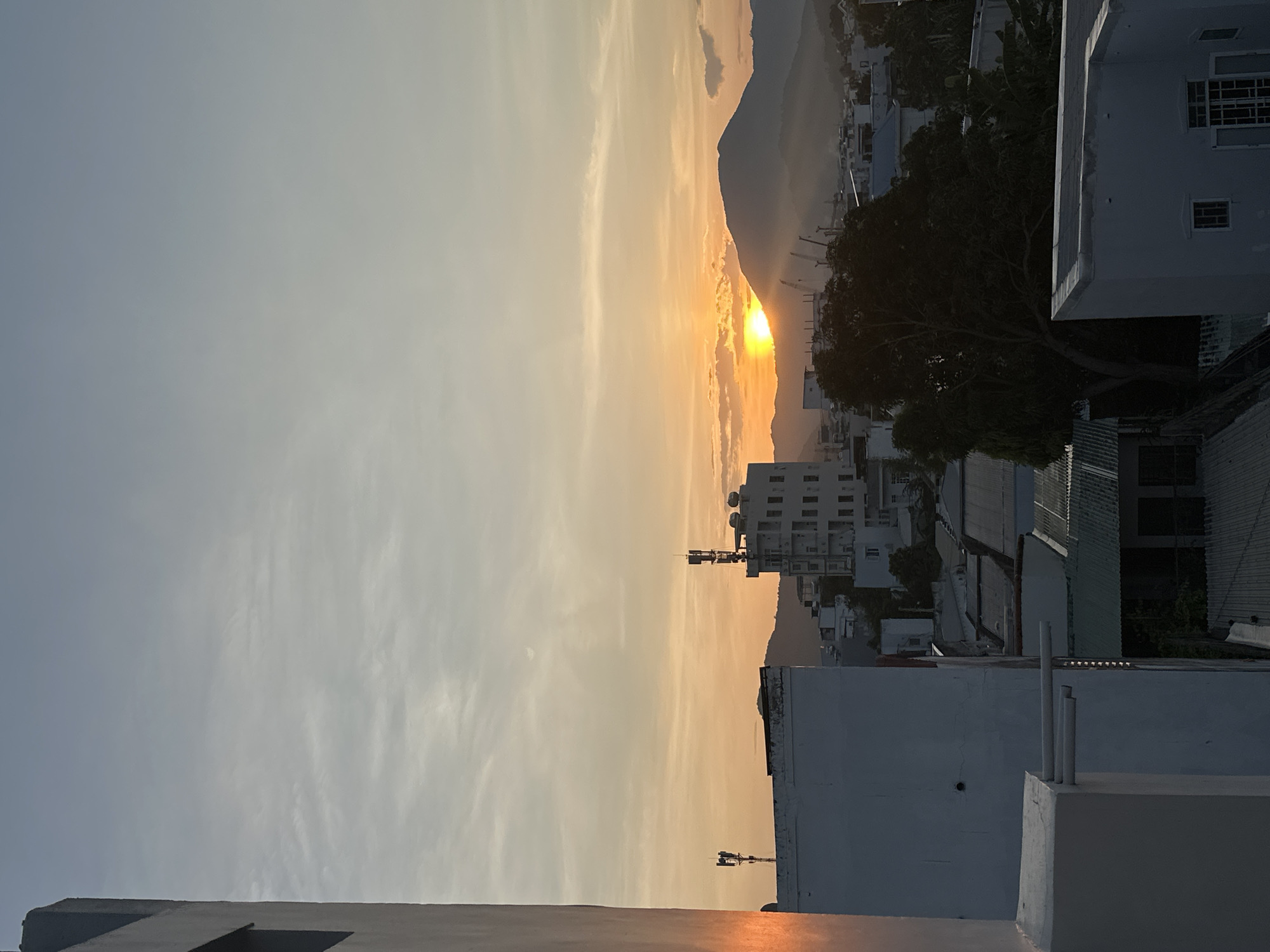 The image depicts a panoramic view of Bac Nha Trang Ward, Vietnam, at either sunrise or sunset, characterized by a low-angle sun casting a warm, golden light across the scene. The sky transitions from a pale blue-grey at the zenith to vibrant orange and yellow hues near the horizon, with thin, scattered clouds, some illuminated by the sun's direct light.
A prominent mountain range is silhouetted against the bright sky in the background, partially obscuring the sun, which appears as an intense, glowing orb. Strong sun rays penetrate through atmospheric haze and cloud layers emanating from behind the mountain, illuminating the air above the city.
The urban landscape in the midground and foreground consists of numerous buildings. On the far left, a tall, light-colored concrete structure with a recessed window frames the left side of the image, its surface reflecting a strong orange glow from the sun. Across the midground, several multi-story buildings with flat, light-colored facades are visible. One taller building towards the center-right features a visible telecommunications mast or antenna array on its rooftop. Another building to the left also has a smaller antenna. Various rooftops of different designs are present, including flat concrete surfaces with equipment such as water tanks, and corrugated metal roofs on lower structures. Distant construction cranes are faintly discernible near the mountain base, suggesting ongoing development. Dark, dense tree canopies are interspersed among the buildings, particularly prominent in the lower central and right portions of the frame, appearing as silhouettes against the brighter background. Atmospheric haze is evident at the base of the mountains and throughout the city, diffusing the light and softening distant details. No individuals are visible within the frame.