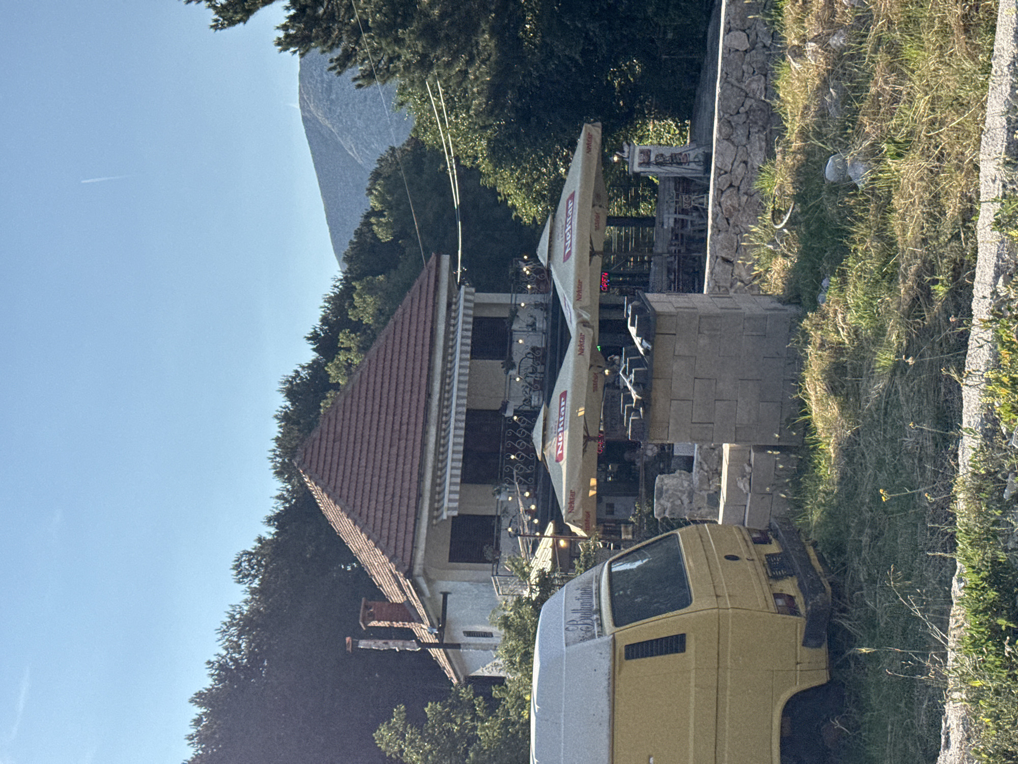 In Njeguši, Montenegro, a yellow camper van is parked on the left side of the image, facing away from the viewer. A house with a red tile roof is visible behind the van, with a balcony overlooking an outdoor seating area. Several umbrellas, branded with "Nektar," are set up over the seating area, suggesting it's a cafe or restaurant. The scene is surrounded by trees, with a mountain range in the background under a clear blue sky. The grounds are a mix of grass and dry vegetation, with some stone walls and structures present.
