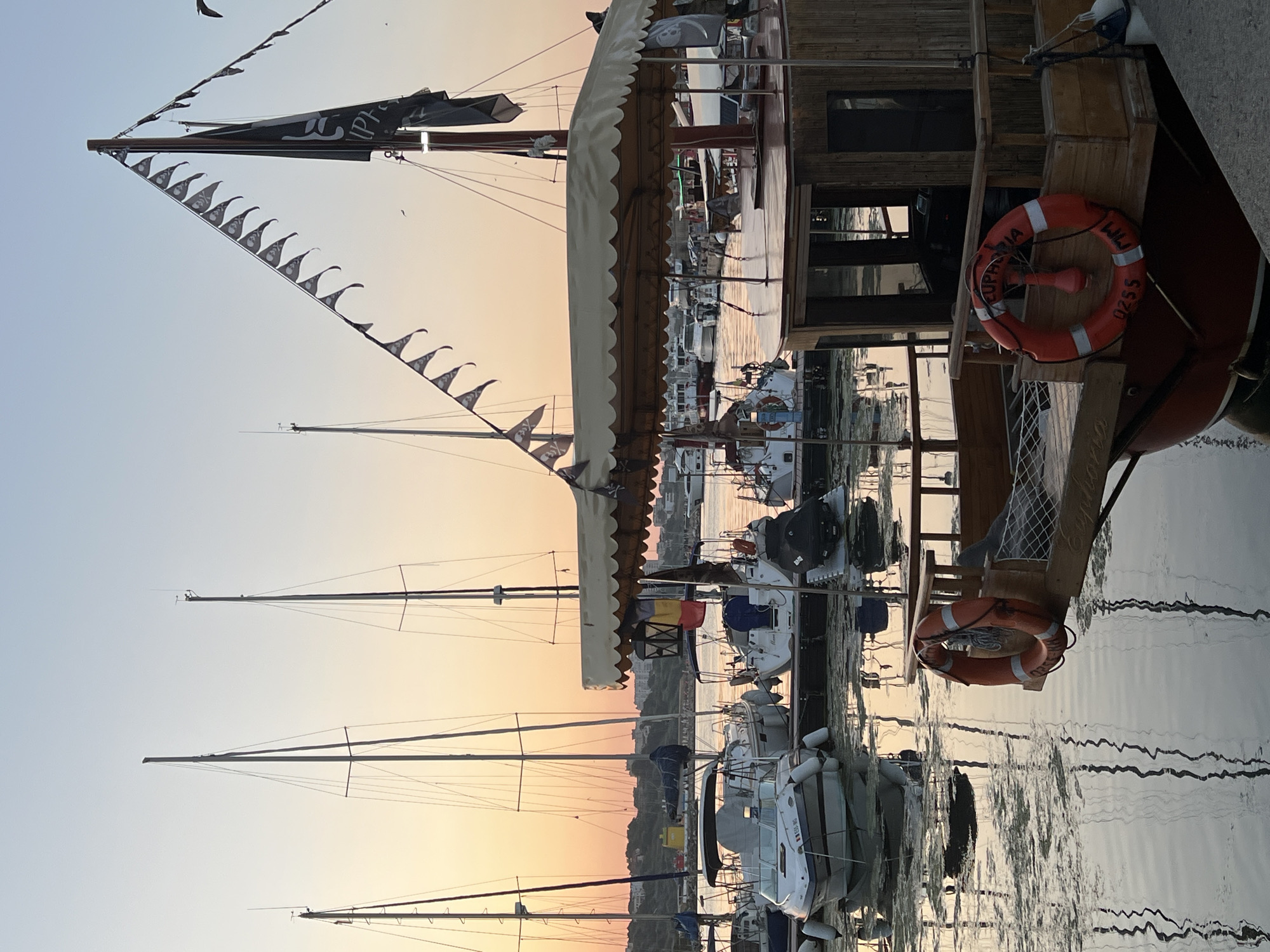 The image depicts a marina at dusk, likely in Constanța, Romania. The sky is a soft gradient of orange and blue, reflecting on the calm water. Several sailboats and smaller motorboats are docked. In the foreground, a wooden boat with a thatched canopy is prominently featured. It has a red life preserver with the word "EUPHORIA" and the number "0255" inscribed on it. A pirate-themed flag and pennant string are visible on its mast. In the background, more boats and buildings along the coastline can be seen. The overall atmosphere is peaceful and serene.