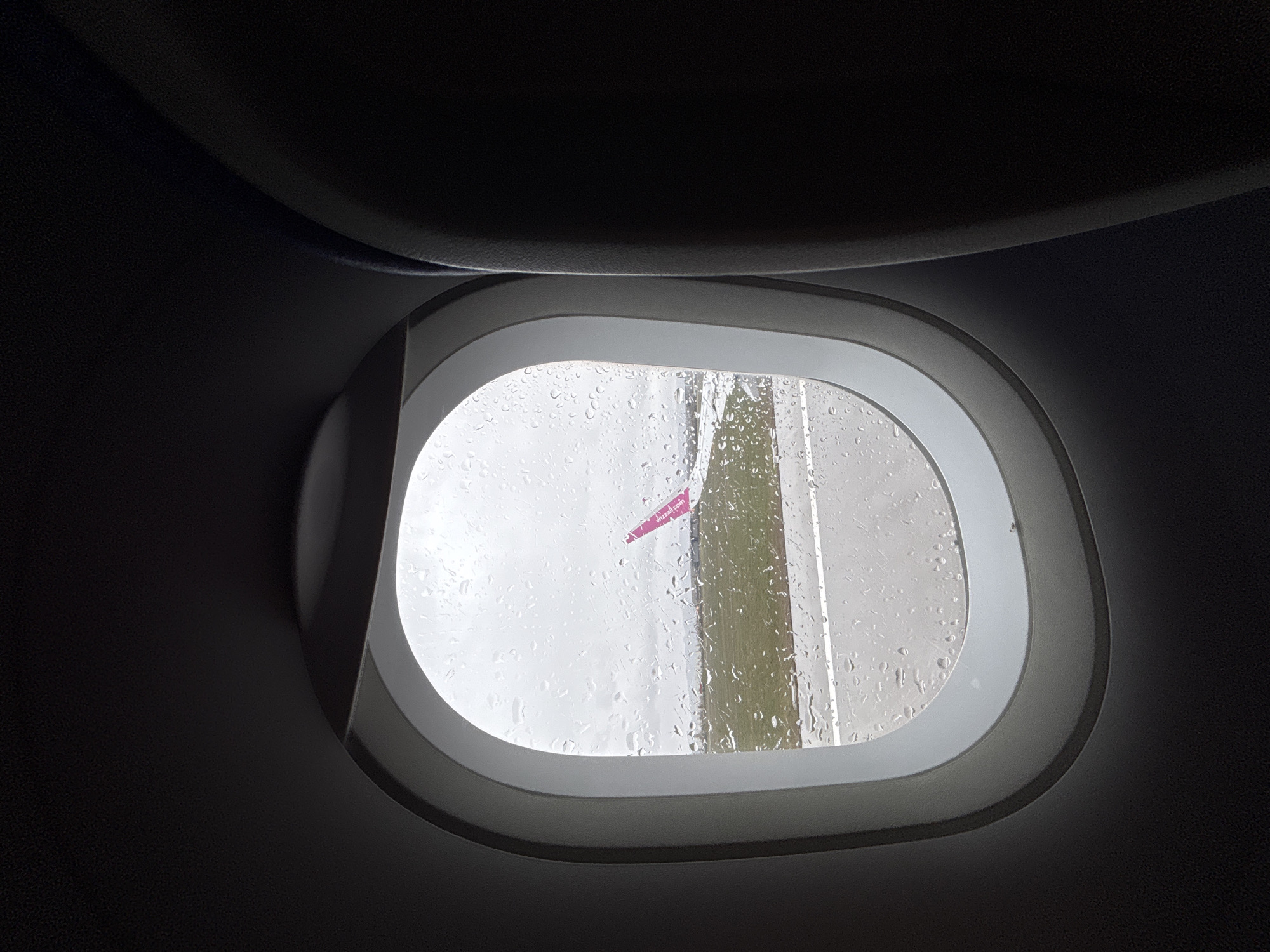 The image shows a view from an airplane window covered in raindrops. Outside the window, part of an airplane wing with the word "wizzair.com" written in magenta is visible. A runway with white lines is also partially visible. The setting appears to be a rainy day. Location is not apparent from the image, the city of Sofia, Bulgaria is not verifiable.