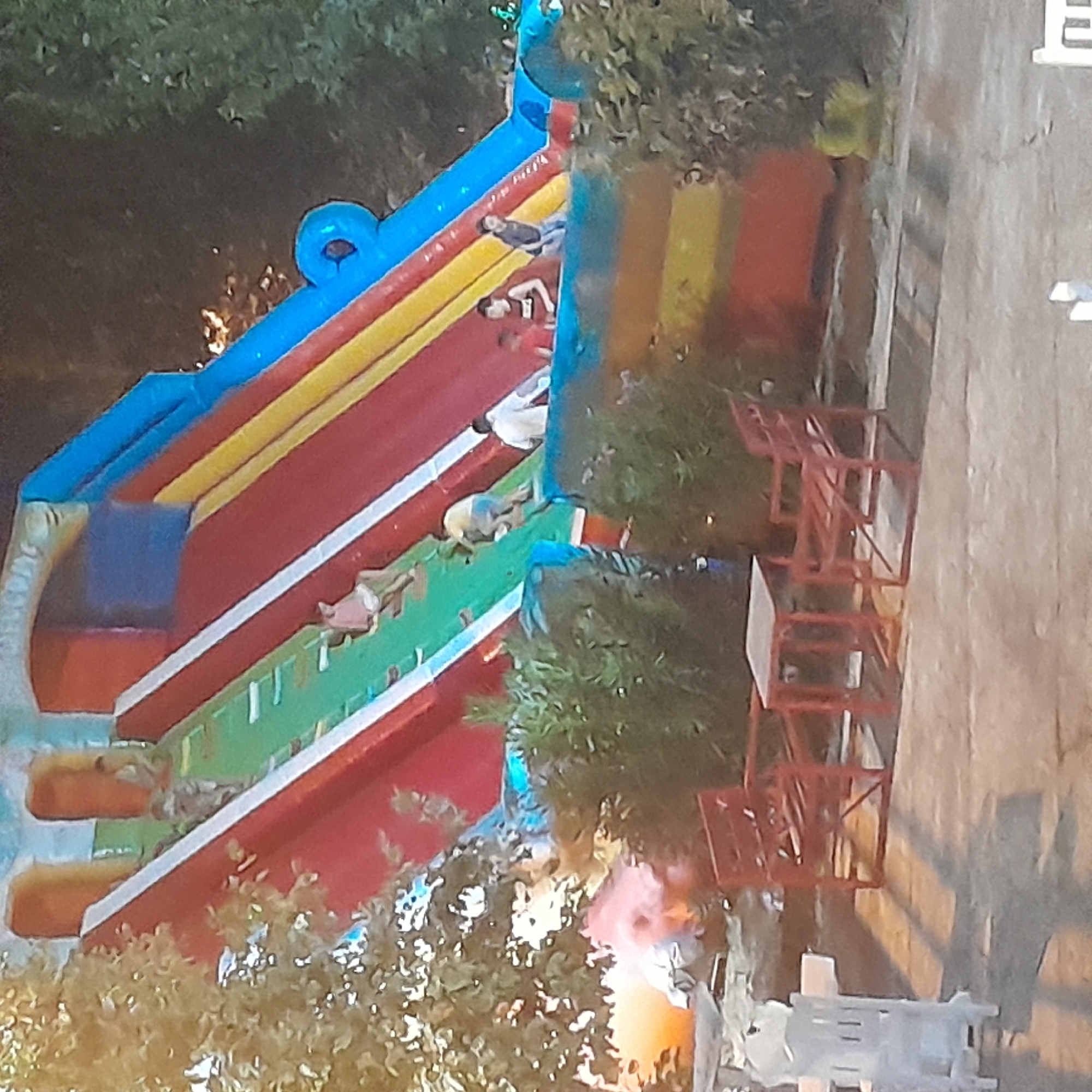 The image shows an inflatable slide with children playing on it. The slide is multi-colored with red, yellow, green, and blue sections. Several children are visible on the slide, some climbing, some sliding. In the foreground, there are red chairs and tables, and a white plastic chair. The setting appears to be outdoors, possibly in a park or recreational area, with trees and bushes visible in the background. The overall scene suggests a lively atmosphere of outdoor recreation. The location is identified as Rudəkənar, Azerbaijan.