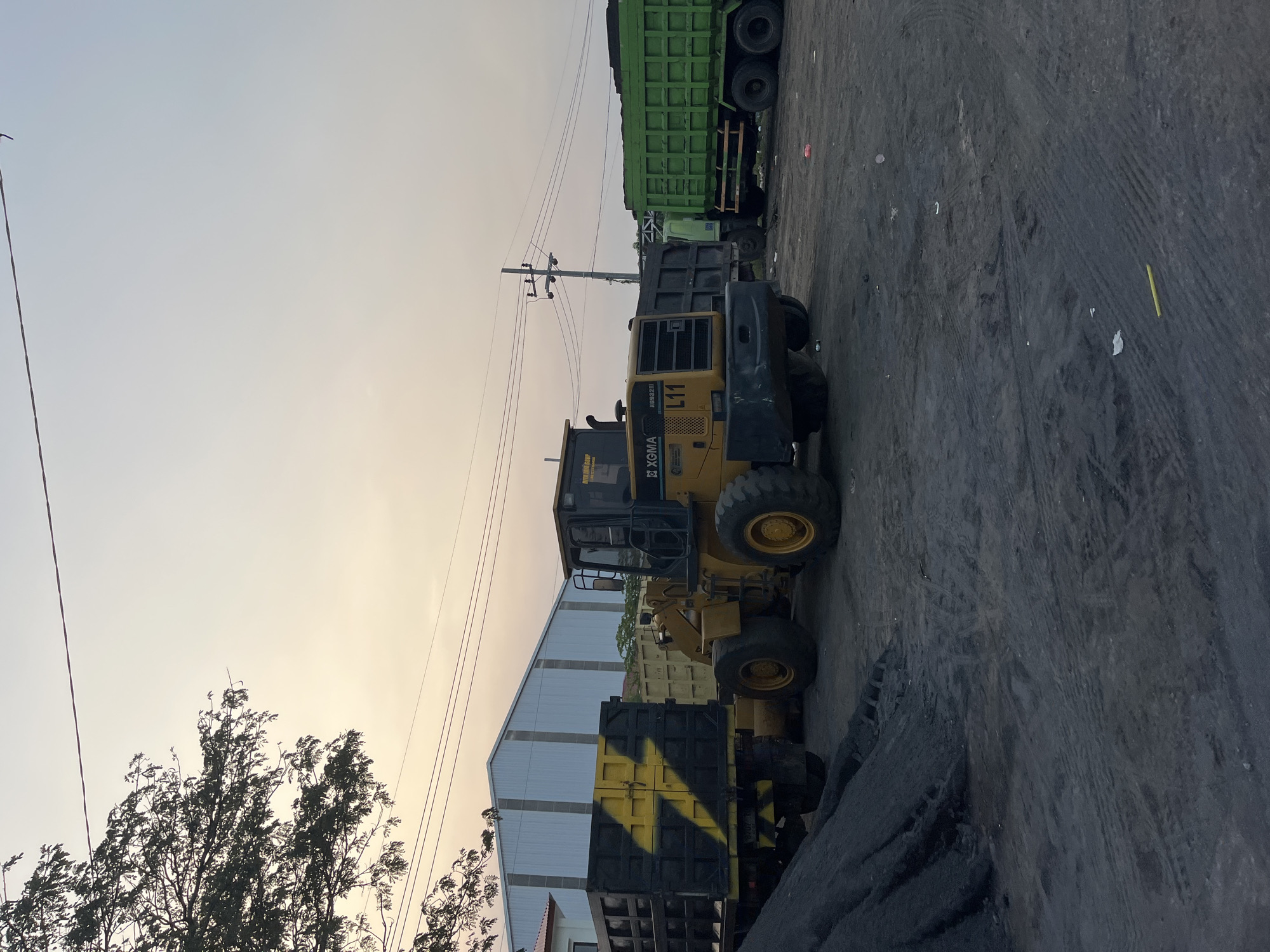 The image depicts an outdoor industrial scene with heavy machinery and trucks under a hazy sky, suggesting either dawn or dusk. In the foreground, a yellow front-end loader with the markings "XGMA" and "L11" is prominent, its tires and bucket visible. Behind it, a truck with a black and yellow striped bed is parked. Further back, a large green dump truck filled with dark material, likely coal, is positioned. A tall utility pole with multiple wires runs across the frame, indicating the presence of infrastructure. The ground is covered in dark, dusty material, possibly coal dust, with tire tracks visible. In the background, a corrugated metal building stands. The overall impression is one of an active industrial site, likely a coal yard or processing facility, possibly in Kedungcangkring, Indonesia, given the context. The lighting suggests a soft, diffused natural light, with no strong shadows, consistent with early morning or late afternoon. No people are visible in the frame.