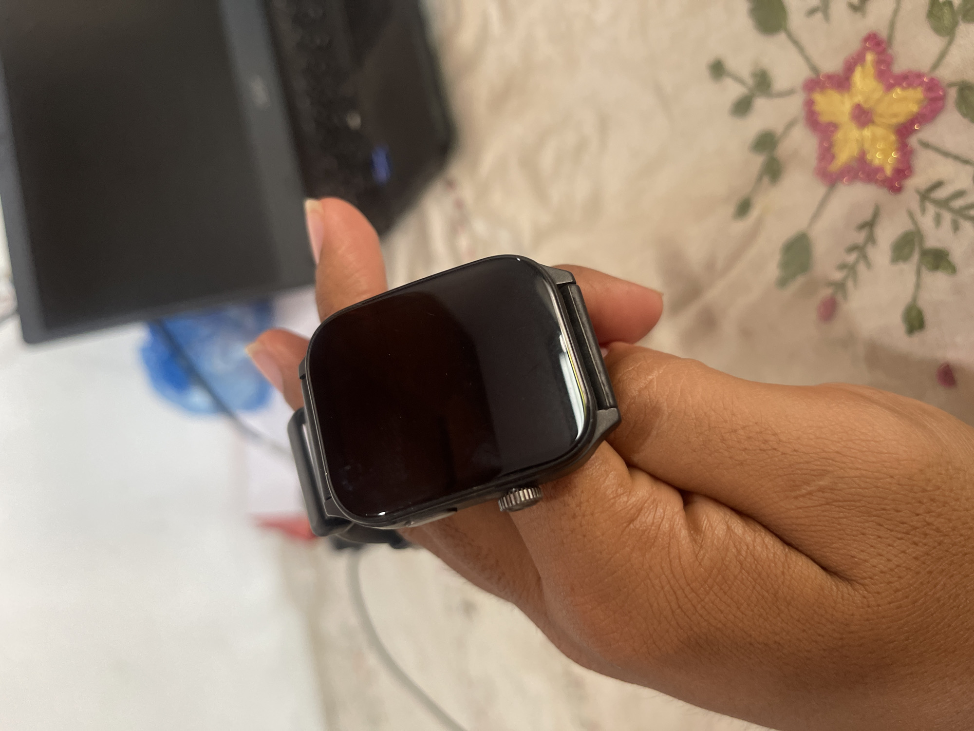 A person's hand is holding a black smartwatch with a square face. The smartwatch has a textured crown on its side. In the background, a laptop is visible, and a floral-patterned cloth is spread out. The scene appears to be indoors. This image was taken in the city of Julicotmeurak, Indonesia.