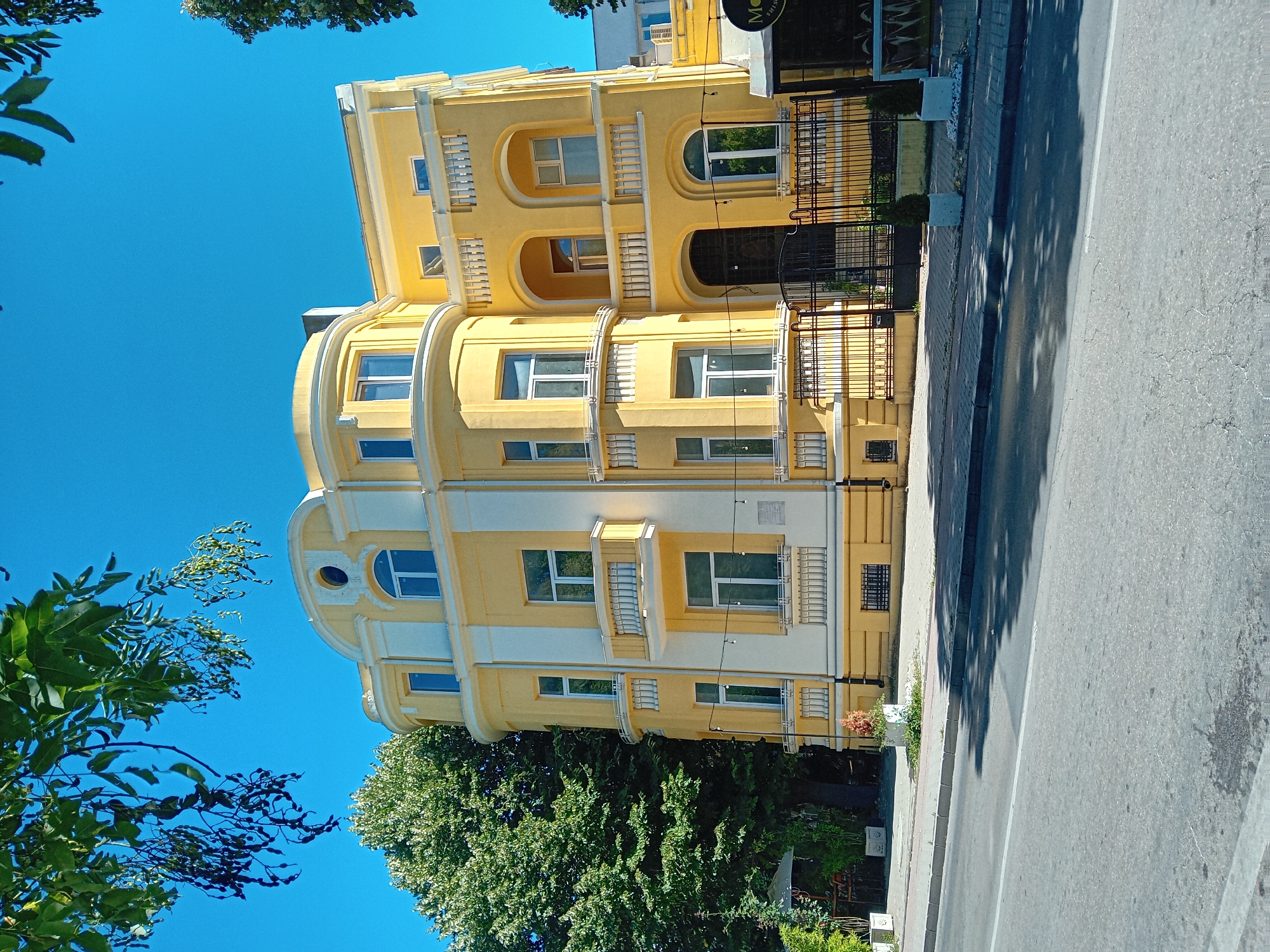 The image shows a bright yellow building with white accents, featuring a curved facade and multiple windows, some with balconies. The building is located in Varna, Bulgaria, on a street with a sidewalk and a road. Tall green trees are visible to the left of the building, and a clear blue sky forms the backdrop. There is a black wrought-iron gate in front of the building. The scene is well-lit, suggesting it was taken on a sunny day.