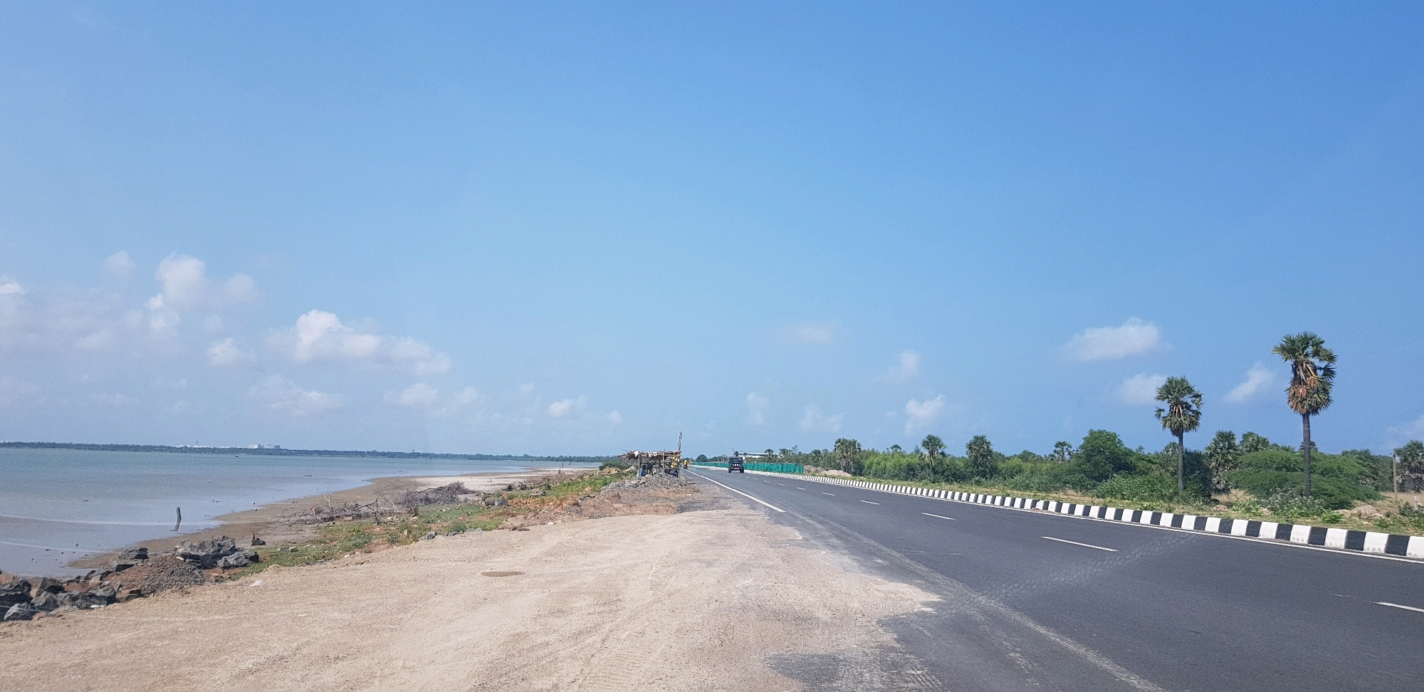 This is a panoramic, eye-level, full shot of a coastal road under a clear blue sky with scattered white clouds. On the left, a calm, shallow body of water meets a sandy shoreline littered with rocks and driftwood. A small wooden post sticks out of the water near the shore. The land on the left transitions to a rough, unpaved area with tire tracks, which then gives way to the paved road on the right. This road, marked with white lines and a black and white striped border, stretches into the distance. On the right side of the road, there is dense green vegetation and several palm trees, with two prominent tall palm trees in the foreground. A black car is visible driving on the road in the middle distance, and some construction or scaffolding is present on the left side of the road further back. The scene appears to be in or near Cheyur, India, given the coastal landscape and vegetation.