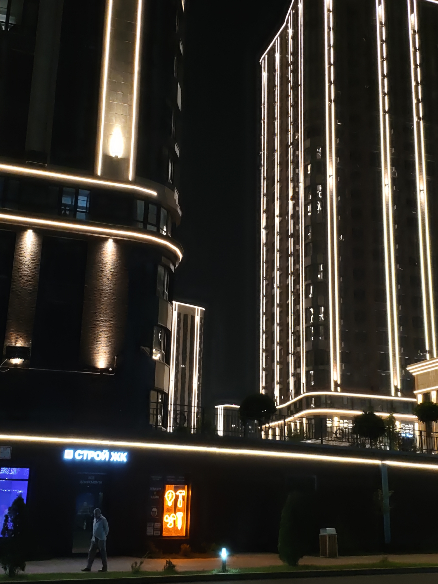 The image depicts a nighttime scene in Rostov-on-Don, Russia, featuring modern architecture with illuminated building facades. Two tall residential buildings, likely apartment complexes, dominate the frame. The building on the left has a darker facade with vertical white LED lighting strips and horizontal lighting along its curved base. The building on the right is similarly lit with vertical strips of light, showcasing a more rigid, rectangular structure. Below these buildings, a lower structure has a dark facade with a prominent illuminated sign reading "СТРОЙ ЖИ" in blue and white letters. To the right of this sign, there is an orange illuminated sign featuring graphic icons of tools and clothing. A solitary man, dressed in light-colored clothing, walks on the sidewalk in front of the lower building. The ground level appears to be a sidewalk with some landscaping, including small trees and bushes. The overall atmosphere is urban and contemporary, with the lighting creating a dramatic and striking visual effect against the dark night sky.