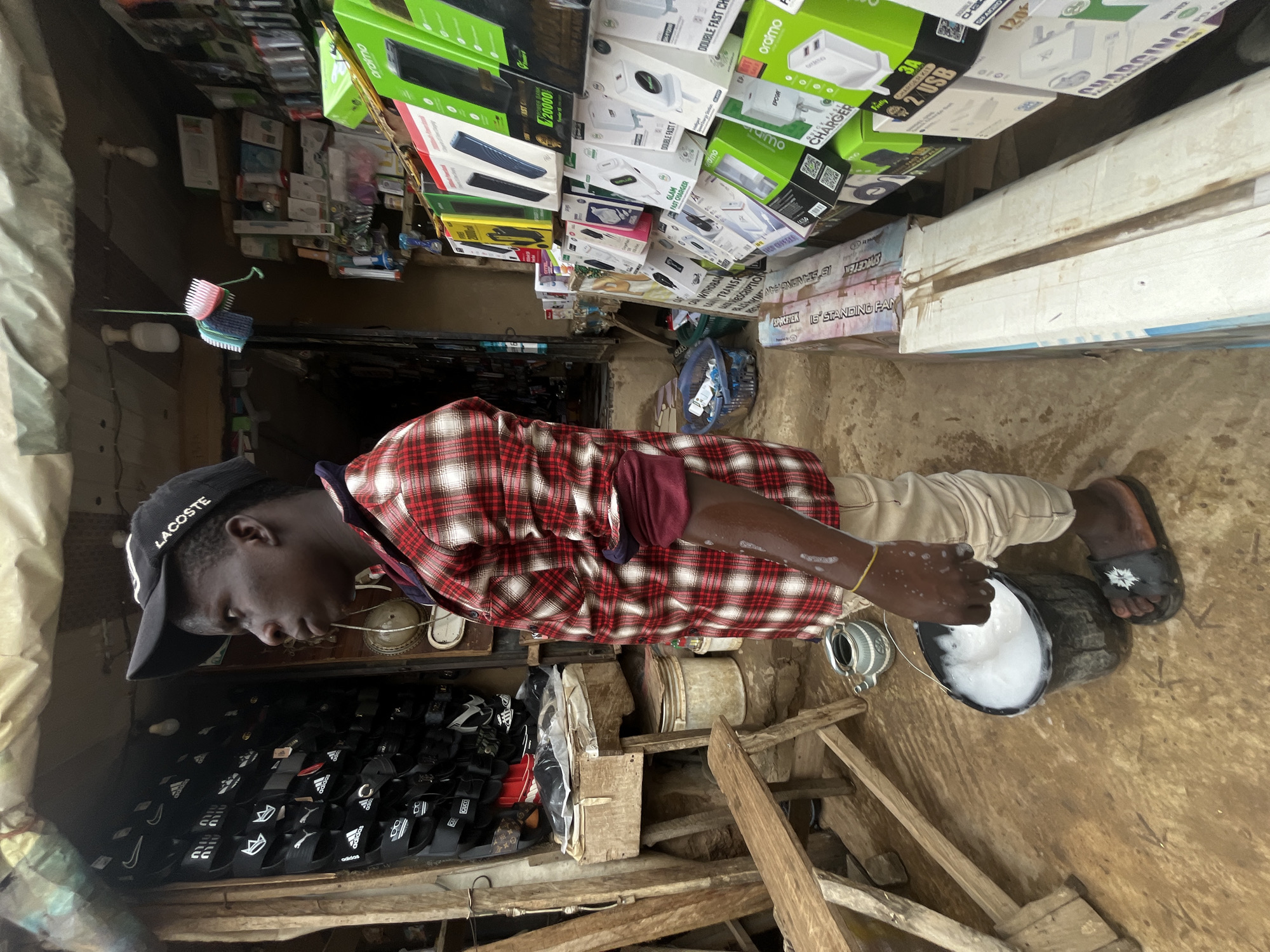 The image depicts a street-side stall in Abuja, Nigeria, with a young man standing near the entrance. He is wearing a red and white plaid shirt, beige pants, and a black baseball cap with "LACOSTE" written on it. His right hand is in a black bucket filled with foamy white liquid, and his arm is wet. To his left, a wooden bench is visible. The stall displays various items, including shelves filled with electronics accessories like phone chargers, power banks, and cables, along with a rack of footwear at the back. A broom hangs from the ceiling. The ground appears to be made of earth and concrete. The overall impression is one of a bustling marketplace environment.