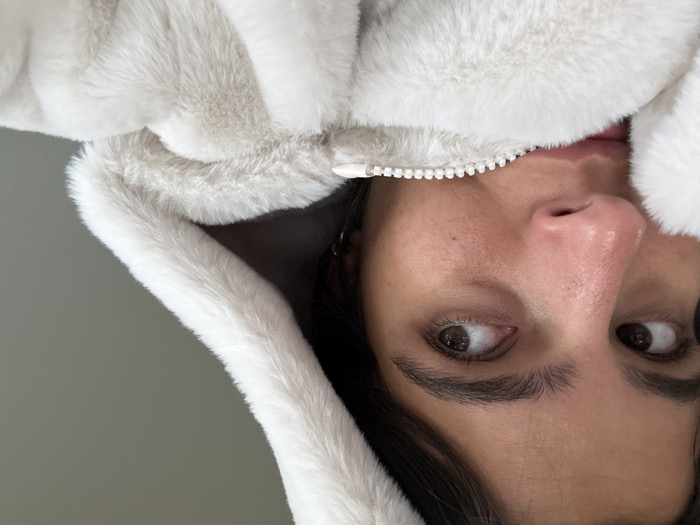 A close-up, eye-level image depicts the upper portion of an individual, likely an adult, in an indoor setting. The subject has dark hair and brown eyes, with visible dark eyebrows. Their gaze is directed slightly to their right (viewer's left). The individual wears a light-colored, likely off-white or cream, faux fur hooded garment. The hood is up and frames the upper right side of their head. The garment's zipper, featuring white plastic teeth, is partially visible and extends upwards, obscuring the subject's mouth and chin. The lower part of the face is covered by the zippered faux fur material. The background is a plain, light grey wall. The scene's specific location within Princeton, United States, is not discernible from the visual evidence.