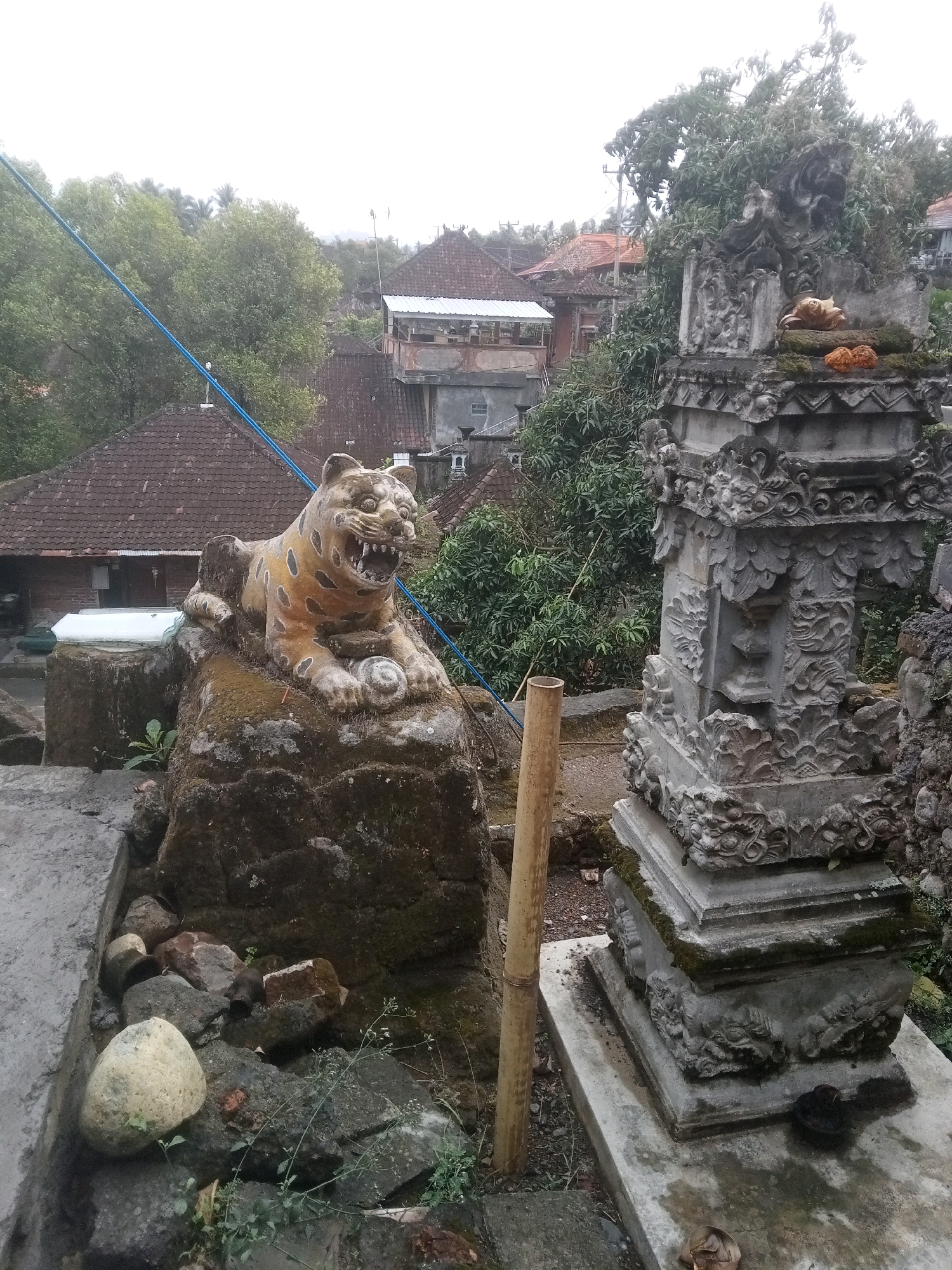 The image depicts an outdoor scene in the city of Mendoyo, Indonesia. In the foreground, a stone sculpture of a tiger with yellow and black markings is positioned on a large, moss-covered rock. The tiger sculpture has its mouth open, revealing teeth, and appears to be holding a coiled object in its paws. To the right of the tiger sculpture is an ornate stone structure, possibly a shrine or altar, adorned with intricate carvings and topped with moss. Two small, orange offerings are placed on the top of this structure. A bamboo pole stands vertically to the left of the stone structure. The background reveals a residential area with traditional Indonesian houses featuring tiled roofs, lush green trees, and a hazy sky, suggesting overcast weather. The ground in the foreground is a mixture of concrete, rocks, and sparse vegetation.