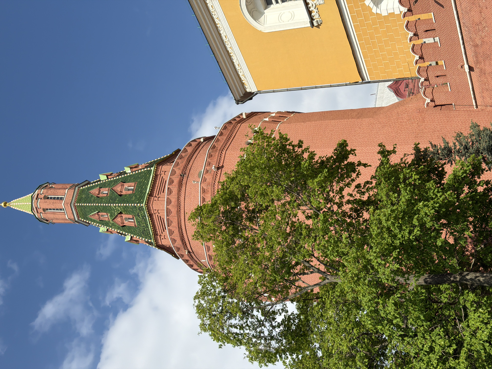 The image depicts a low-angle view of the Kremlin's Trinity Tower in Moscow, Russia. The tower, made of red brick with green tile accents on the spire, is partially obscured by a large tree with green foliage. The sky is visible, with blue hues and scattered white clouds. A yellow building is seen in the upper right corner of the image.