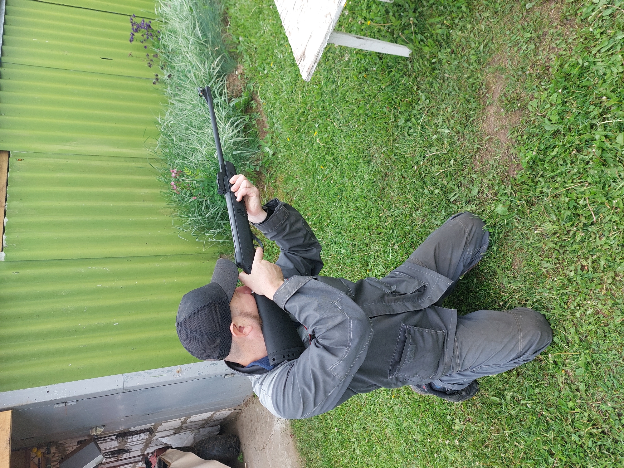 An adult male is observed kneeling in a grassy outdoor area in Большой Букор, Russia, aiming a black rifle. He wears a black baseball cap, a grey long-sleeved work jacket, and grey trousers. His right knee is on the ground, and his left knee is bent upwards. The rifle's buttstock is pressed against his right shoulder, and his head is turned to align his right eye with the rifle's sight. His left hand supports the rifle's fore-end, and his right hand is on the grip and trigger area. A small puff of vapor or smoke is visible emanating from the rifle's muzzle. In the background, a light green corrugated metal fence extends horizontally. To the left of the fence is a grey building, possibly a shed, with an open doorway and a tire visible near its base. To the right of the man, a rustic white wooden table with two visible legs is present. The ground is covered with green grass, interspersed with weeds and some patches of bare earth. Taller grasses and flowering plants, including purple and pink varieties, grow along the base of the green fence.