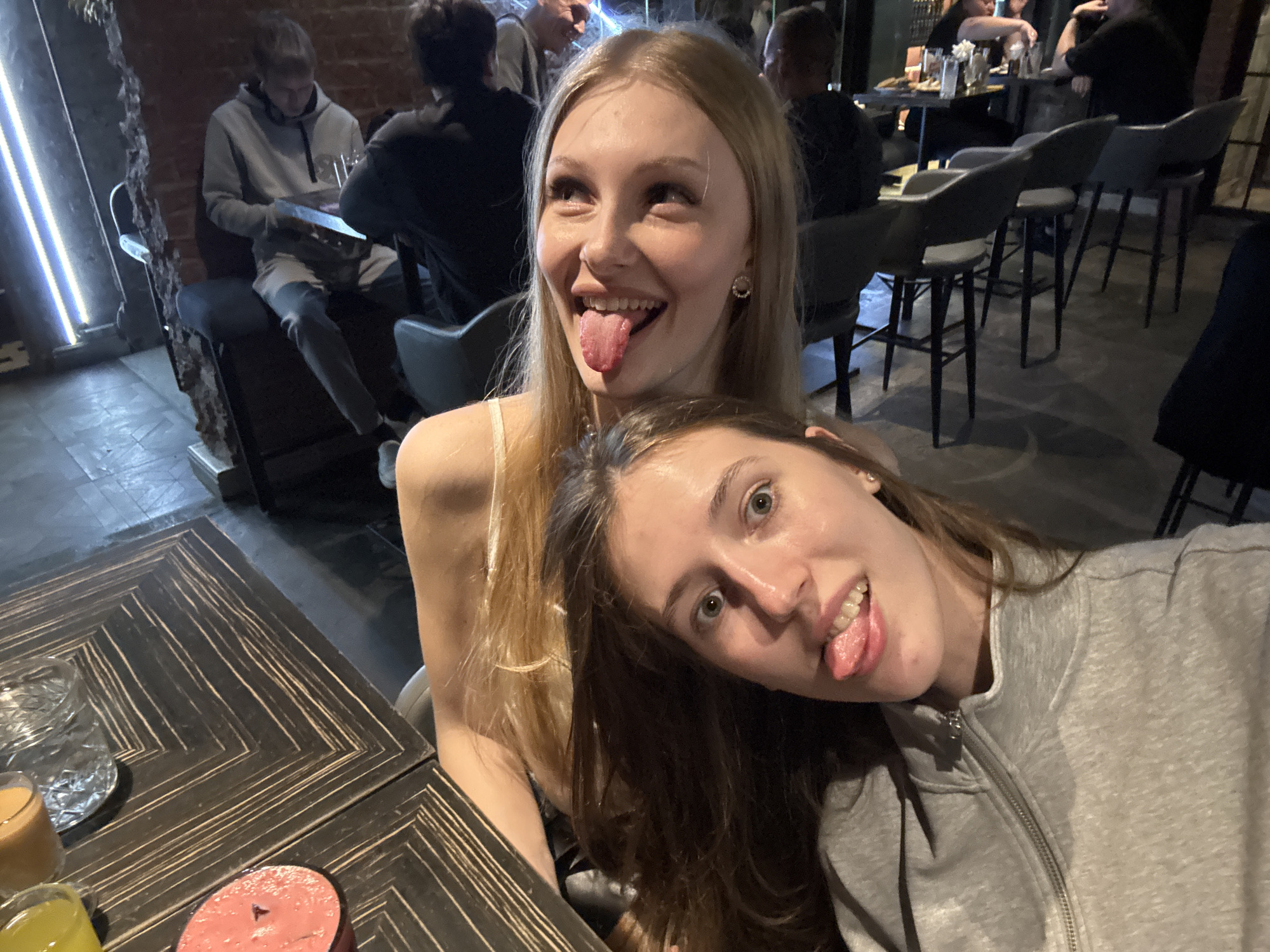 This media file shows two young women in a dimly lit bar or restaurant, likely in Perm, Russia. They are taking a selfie, with one woman in the foreground and another behind her. Both women have their tongues sticking out, suggesting a playful and lighthearted mood. The woman in the foreground is wearing a grey zip-up hoodie, and the woman behind her has blonde hair and is wearing a light-colored top.
The background shows other patrons at tables and bar stools, indicating a social gathering. There are drinks on a wooden table in the foreground, including a glass with ice and two other drinks in smaller glasses. The overall ambiance appears to be casual and informal. The lighting is dim, with some neon lights visible in the background, suggesting it is nighttime. There is no visible text in the image. The women's expressions and the selfie pose convey a sense of fun and camaraderie.