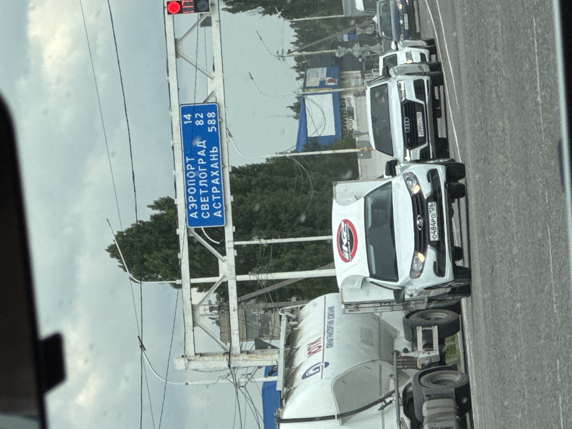 This image shows a street scene in Rozhdestvenskaya, Russia. The perspective is from inside a vehicle, looking out. The primary focus is a blue road sign indicating distances to various locations. The sign reads "АЭРОПОРТ" (Airport) with a distance of "14", "СВЕТЛОГРАД" (Svetlograd) with a distance of "82", and "АСТРАХАНЬ" (Astrakhan) with a distance of "588". The sign is mounted on a white metal structure, possibly an overpass or gantry.
In the background, there are several vehicles on a road, including cars and trucks. One white truck, possibly a cement mixer, is prominent on the right side of the frame. Another white vehicle, an SUV, is also visible. The sky is overcast, suggesting a cloudy day. Green trees line the side of the road in the distance. A traffic light with a red signal is visible at the top left of the image. The overall scene appears to be during daylight hours. The road surface is asphalt. There is no clear indication of people within the vehicles. The image has a slightly grainy quality, and the overall lighting is diffused due to the cloud cover.