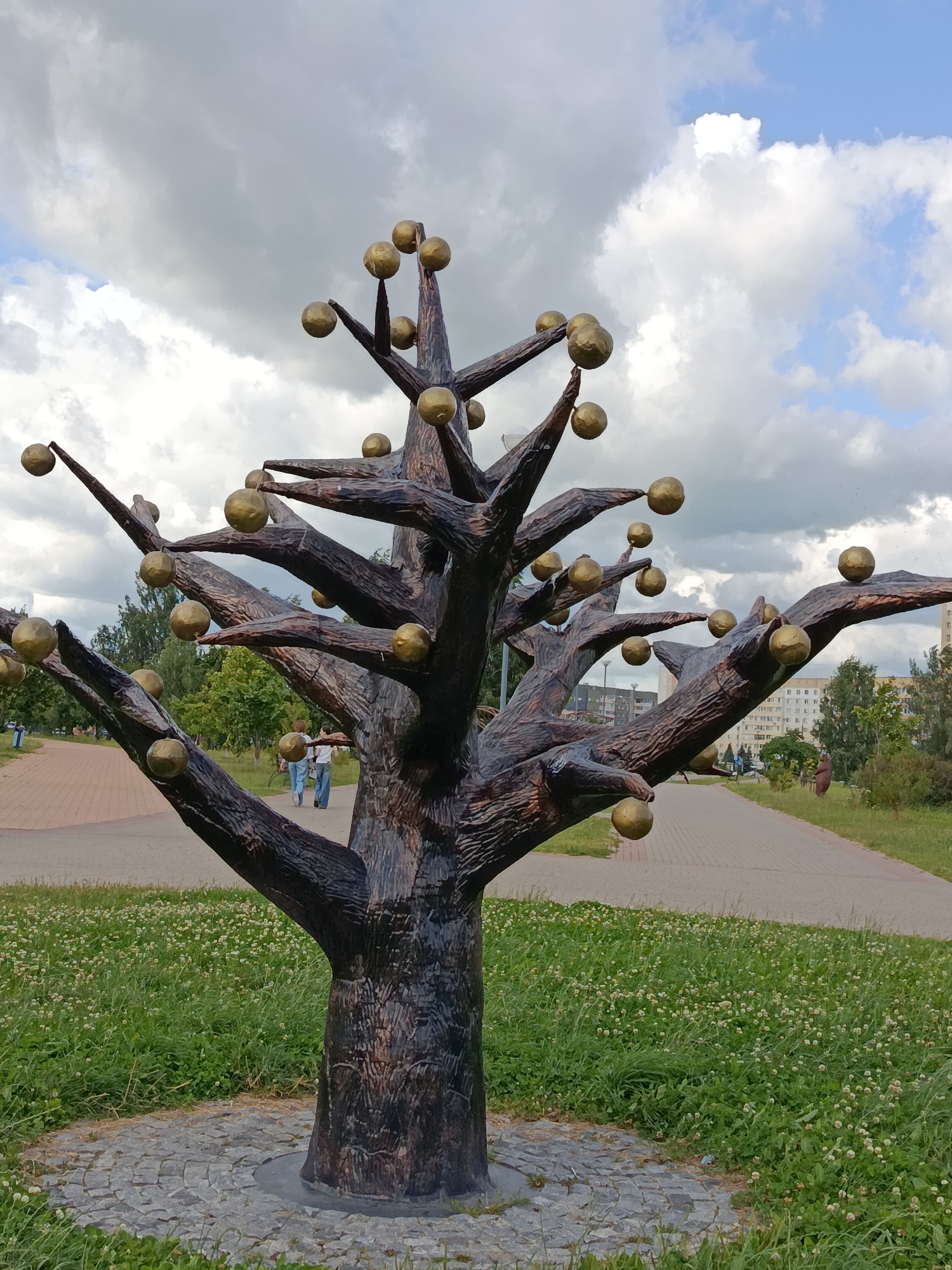 This media file shows a bronze sculpture of a tree with golden spheres for fruit, located in a park in Zhlobin, Belarus. The sculpture is the central subject, with its textured, dark bronze trunk and branches spread wide. Golden spheres, resembling apples or orbs, are attached to the ends of many branches. The tree is situated on a circular cobblestone base, surrounded by lush green grass dotted with small white flowers.
In the background, a paved pathway winds through the park. Two people, a couple, are walking along this path, their backs to the camera. They appear to be casually strolling. Further in the distance, more people can be seen walking, and a bicycle is visible. Trees and some low-rise buildings are present in the background, suggesting an urban park setting.
The sky is partially cloudy with patches of blue sky peeking through. The lighting suggests it is daytime, likely late morning or afternoon. The overall atmosphere is peaceful and relaxed, indicative of a pleasant day outdoors in a park. There is no visible text in the image.