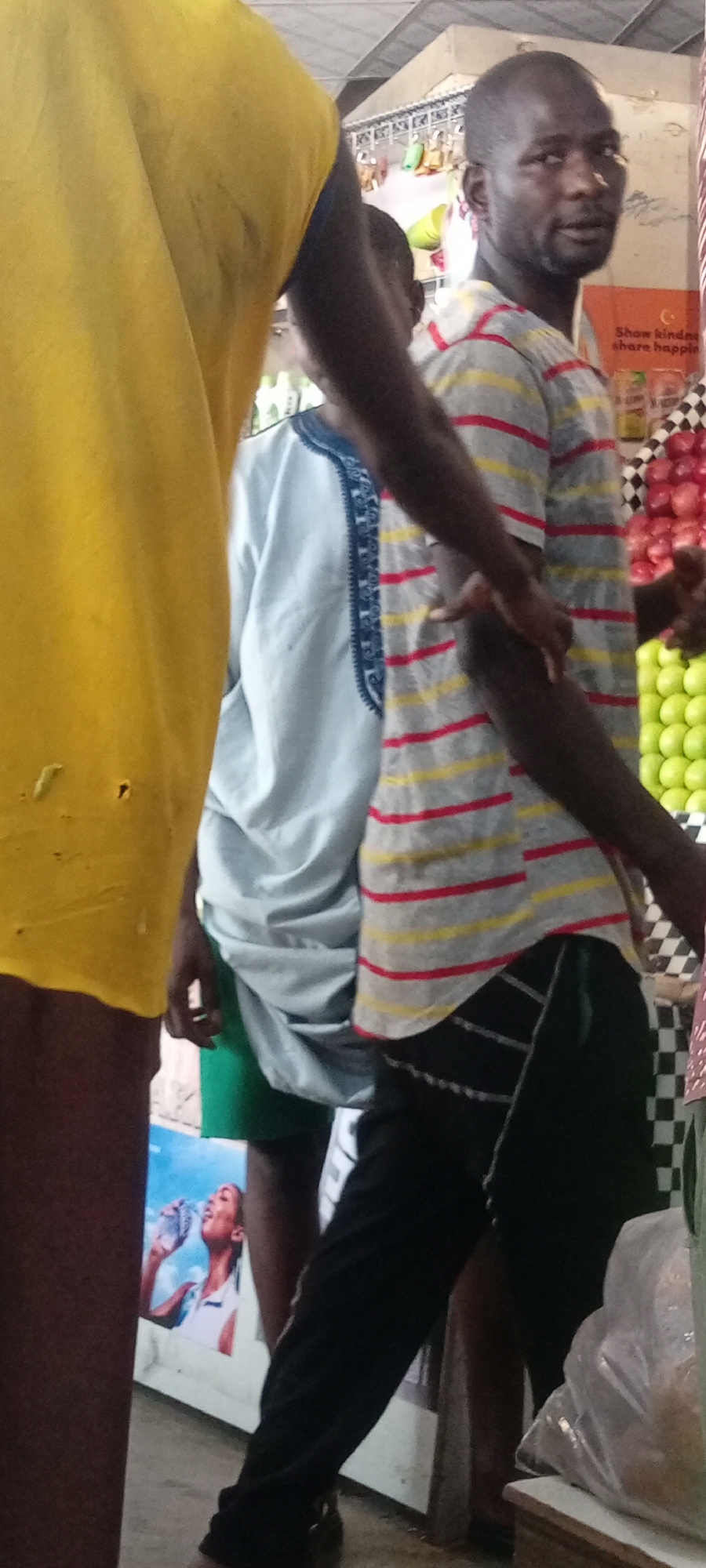 A crowded outdoor market scene in Aviation Quarters, Nigeria, with multiple individuals. A man in a gray and striped t-shirt and black pants looks to the right, partially obscured by the arm and back of a person in a yellow sleeveless shirt. A third individual is visible in a light blue shirt and green shorts. In the background are displays of fruits (apples, limes), and an advertisement featuring a woman drinking from a water bottle. A checkered tile design can also be seen in the background.