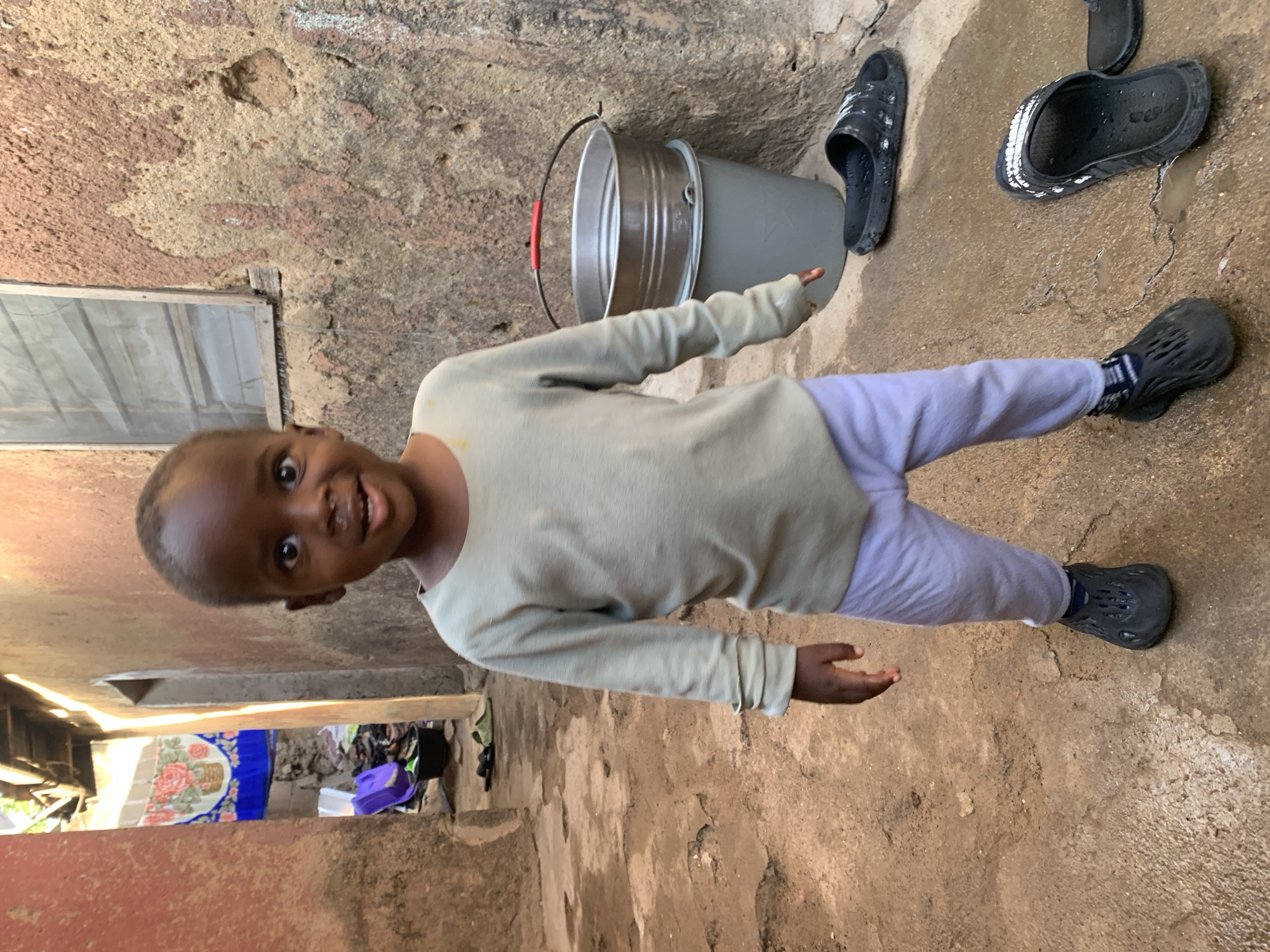 A young child stands on a textured, slightly damp concrete or earthen floor in what appears to be an outdoor courtyard or alleyway. The child wears a light-colored long-sleeved shirt, light purple pants, and black clog-style shoes. To the right of the child are two pairs of wet, black sandals. Behind the child is a light gray bucket with a red handle accent. The walls are rough, uneven, and stained, suggesting weathering and age. In the background, there's a glimpse into a darker interior space with a patterned textile or rug visible. Given the reference point of Jos, Nigeria, it is reasonable to infer this location is within the city's residential or communal areas.