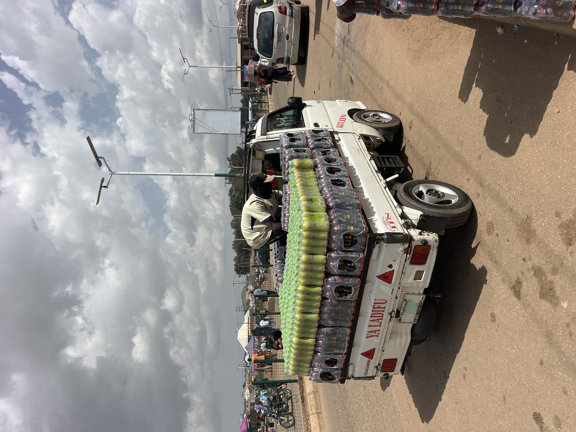 An outdoor shot taken from a low angle shows a white pickup truck heavily laden with drinks. The truck is parked on a paved road, and its sides are stacked with rows of plastic bottles of yellow liquid, shrink-wrapped together. Below these are rows of dark bottles, also shrink-wrapped. The truck has "YA LADIFU" printed in red letters on its side, and a license plate that reads "PLATEAU TOYOTA". A man in a light-colored shirt and a white face mask is leaning over the back of the truck, seemingly adjusting the load. The background is blurred but suggests a busy marketplace with tents, people, a motorcycle, and other vehicles. The sky above is cloudy, with patches of blue sky peeking through.