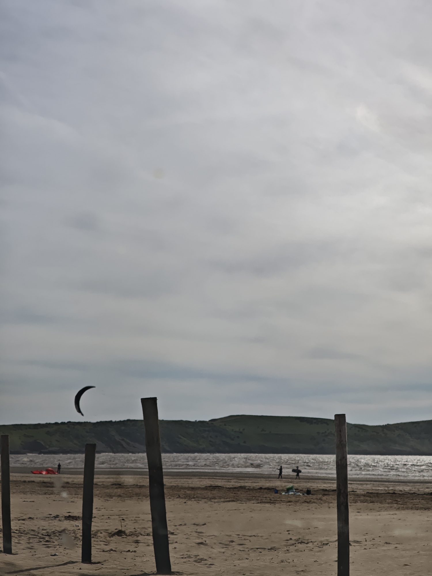 This image depicts a beach scene in Weston-super-Mare, United Kingdom. In the foreground, there are several dark, wooden posts vertically positioned in the sand. The beach is sandy, with visible footprints and other markings. In the midground, there is a body of water (possibly the sea) with waves, and beyond that, a green, hilly landscape. There are a few indistinct figures on the beach near the water. A kite is flying in the sky. The sky is overcast with gray clouds. A red object is to the left of the kite.