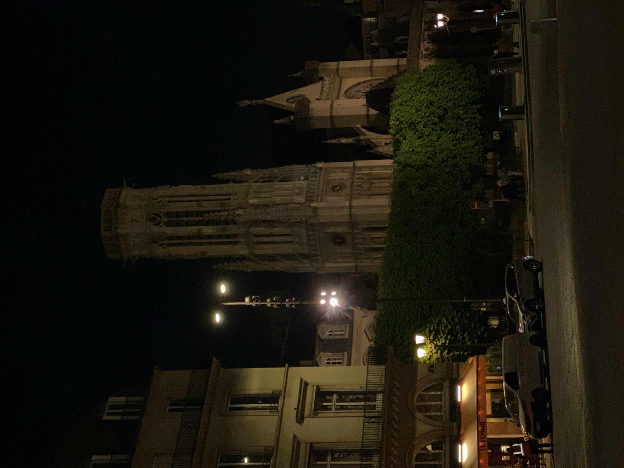 The image captures a nighttime view of a street scene in what appears to be an urban setting, likely Paris, France, given the architectural style. The dominant feature is a tall, ornate Gothic-style tower, possibly resembling the Tour Saint-Jacques, standing prominently against a dark, clear night sky on the left side of the frame. This tower exhibits intricate stonework, arched windows, and decorative elements including visible gargoyles near its upper sections.
To the right of the tower, a dense line of green trees or bushes partially obscures another large stone building with more Gothic architectural details, including what looks like a rose window or circular motif. Further to the right, a multi-lane street is visible, bordered by sidewalks. Several modern streetlights with multiple fixtures illuminate parts of the scene, casting light on the buildings and street.
Along the street, several vehicles are parked, including a dark sedan and a white utility van. On the far right, there are more buildings, one of which has a reddish-orange awning or sign with faint, illuminated text that appears to read "LE FIL MOND" or similar. A single person is dimly visible near the parked van and the building with the sign. The scene conveys the quiet ambiance of a city street after dark, with artificial lights providing the main illumination. The overall impression is one of historical architecture coexisting with contemporary urban life.