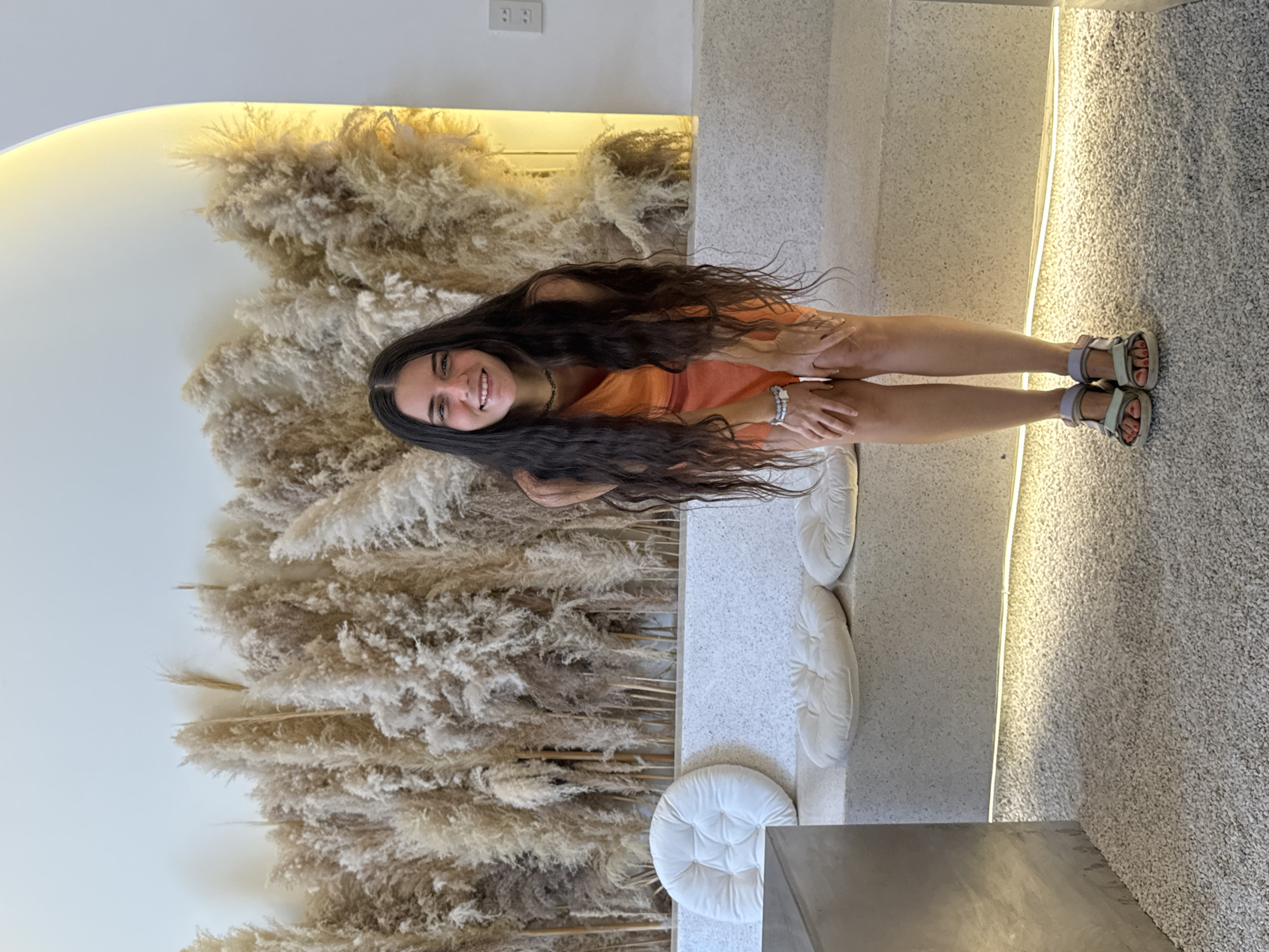A young woman with long, dark hair, wearing an orange top and sandals, is captured in an indoor setting in Phu Thuy Ward, Vietnam. She is smiling broadly at the camera, with a slightly crouched or leaning posture.
Behind her is a prominent wall feature composed of dried pampas grass or similar reeds, creating a soft, textured, and natural backdrop. To her right, a light-colored, speckled bench or low wall is visible, adorned with several white, circular, tufted cushions. A thin strip of warm LED lighting runs along the edge of this structure, casting a gentle glow. The floor to the right is covered with a light-toned, small-grained material, possibly gravel-textured flooring or a short-pile carpet.
In the upper background, above the pampas grass, two white electrical outlets are mounted on a plain white wall. A low, gray surface, possibly a concrete table or bench, is partially visible in the foreground. The overall ambiance suggests a modern, minimalist space, likely a cafe, restaurant, or lounge, characterized by natural elements and contemporary design. The bright lighting suggests it is daytime, enhanced by internal light sources. The woman's cheerful expression conveys a sense of happiness. No discernible text is visible in the image.