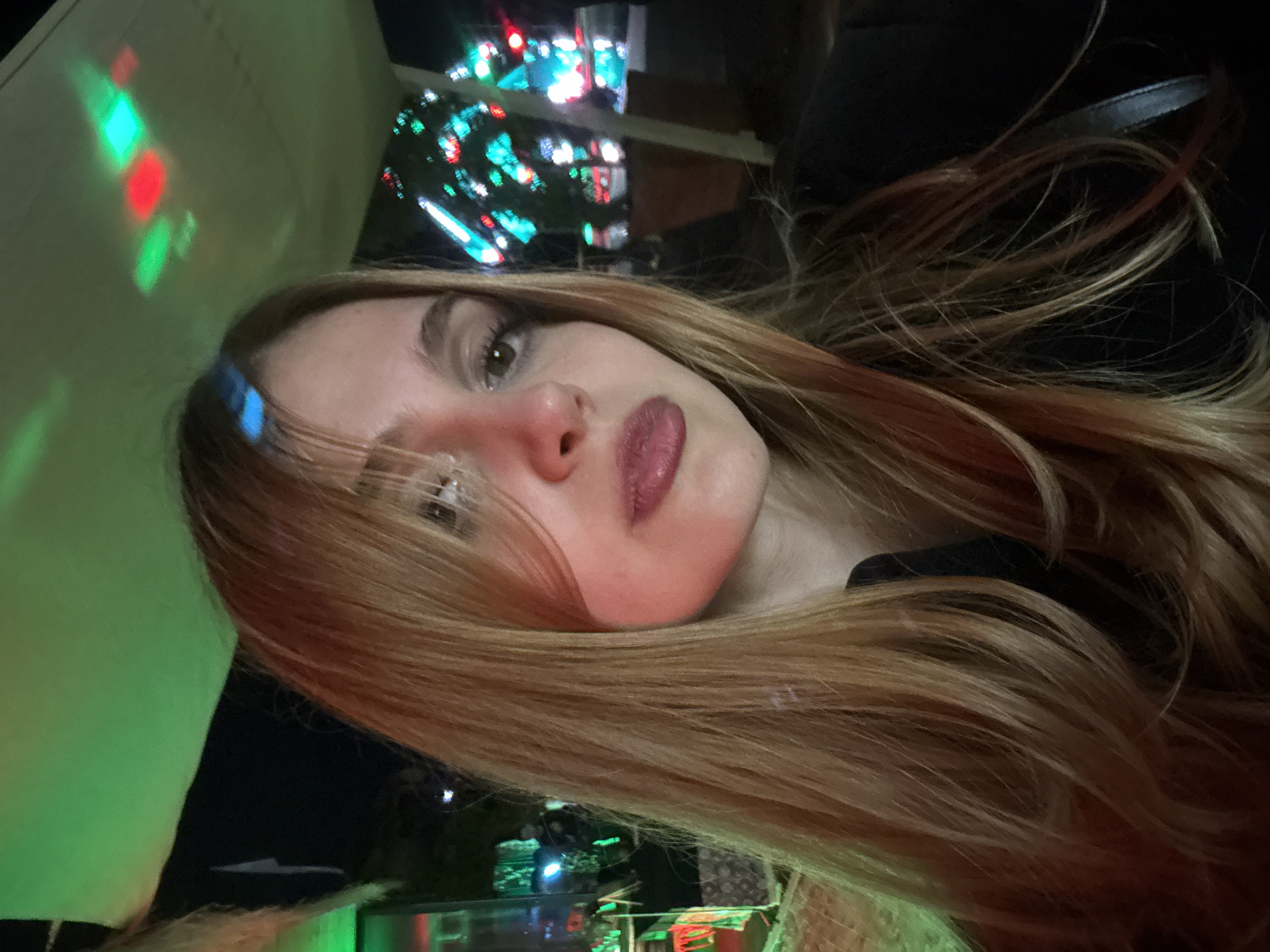 A selfie taken indoors, rotated counter-clockwise approximately 90 degrees. A woman with long, light brown hair and fair skin looks directly at the camera. Her lips are colored a dark shade of pink. Red and green lights illuminate her face. In the background, partially visible, is a blurred scene with numerous small, bright lights, possibly an urban setting or festive lighting. The scene appears to be in Parakar, Armenia based on additional information from the photographer. No specific actions or interactions can be determined from the still image. The environment appears to be dimly lit with artificial, colored light sources.