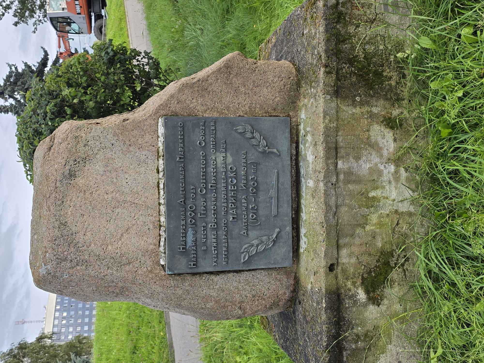 The image shows a stone monument with a metal plaque attached to its front. The plaque bears Russian inscriptions and a depiction of a ship. The monument is situated outdoors, on a concrete base, surrounded by grass and foliage. In the background, a building, trees, and an orange construction vehicle are visible. The overall scene suggests a memorial or historical marker. The inscription on the plaque translates to "Embankment named after Alexander Marinesko. Named in 1990 in honor of the Hero of the Soviet Union, participant of the East Prussian operation, legendary submariner Marinesko Alexander Ivanovich (1913-1963)." This monument is located on the bank of the Pregolya River in Kaliningrad, Russia, dedicated to the Soviet submarine captain Alexander Marinesko.
